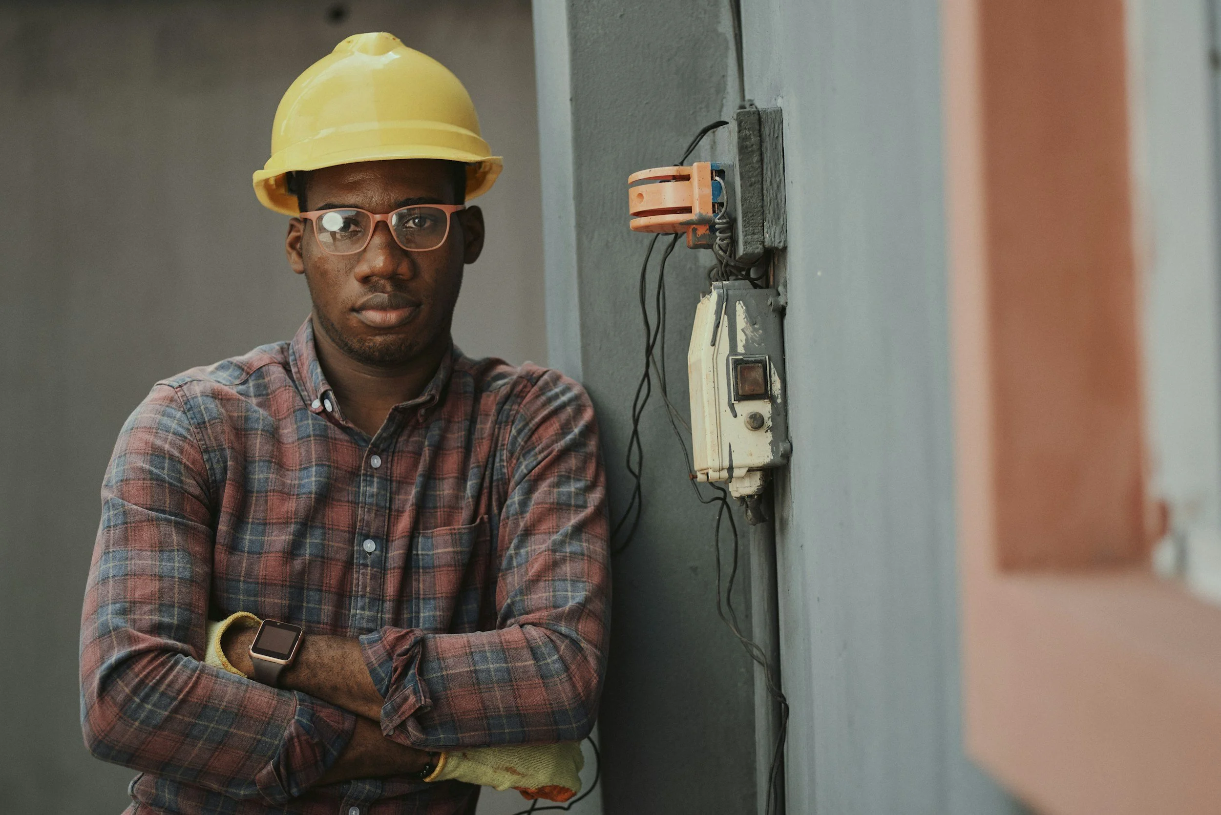 A man wearing glasses, a yellow hard hat, and a plaid shirt stands with arms crossed next to an electrical panel or control box mounted on a gray wall.