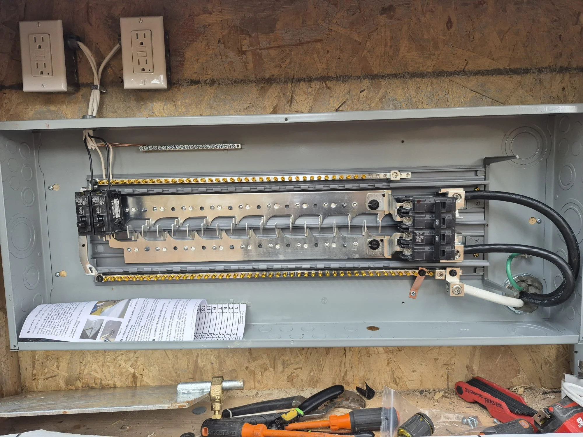 Electrical breaker panel with wiring and circuit breakers inside a gray metal box, mounted on a wooden surface.