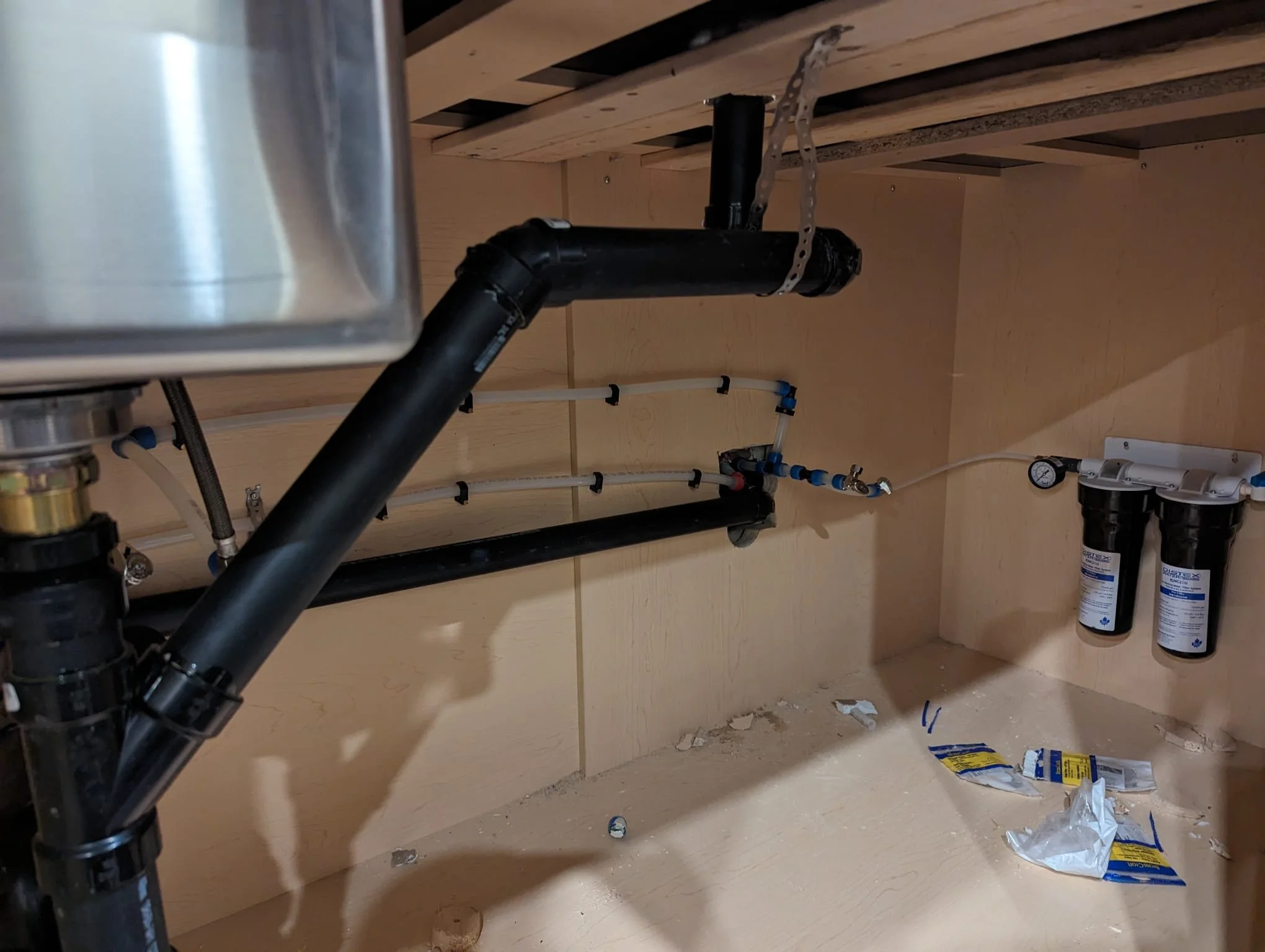 Under-sink plumbing with black pipes, water filtration system with two filters, and a water pressure gauge in a wooden cabinet, with some debris and packaging on the floor.