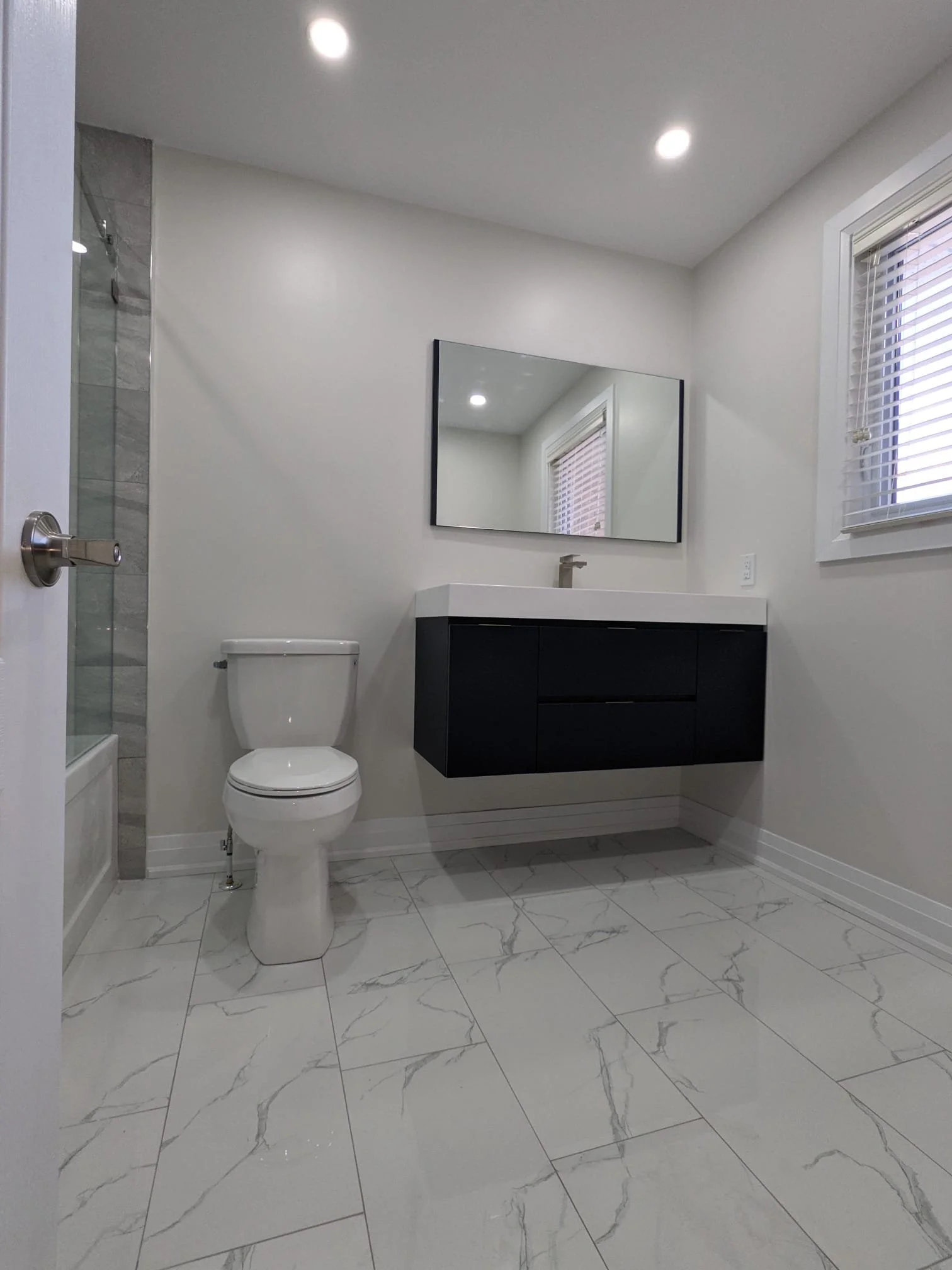 Modern bathroom with a white toilet, black floating vanity with a white sink, large mirror, marble flooring, window with blinds, and a glass-enclosed shower.