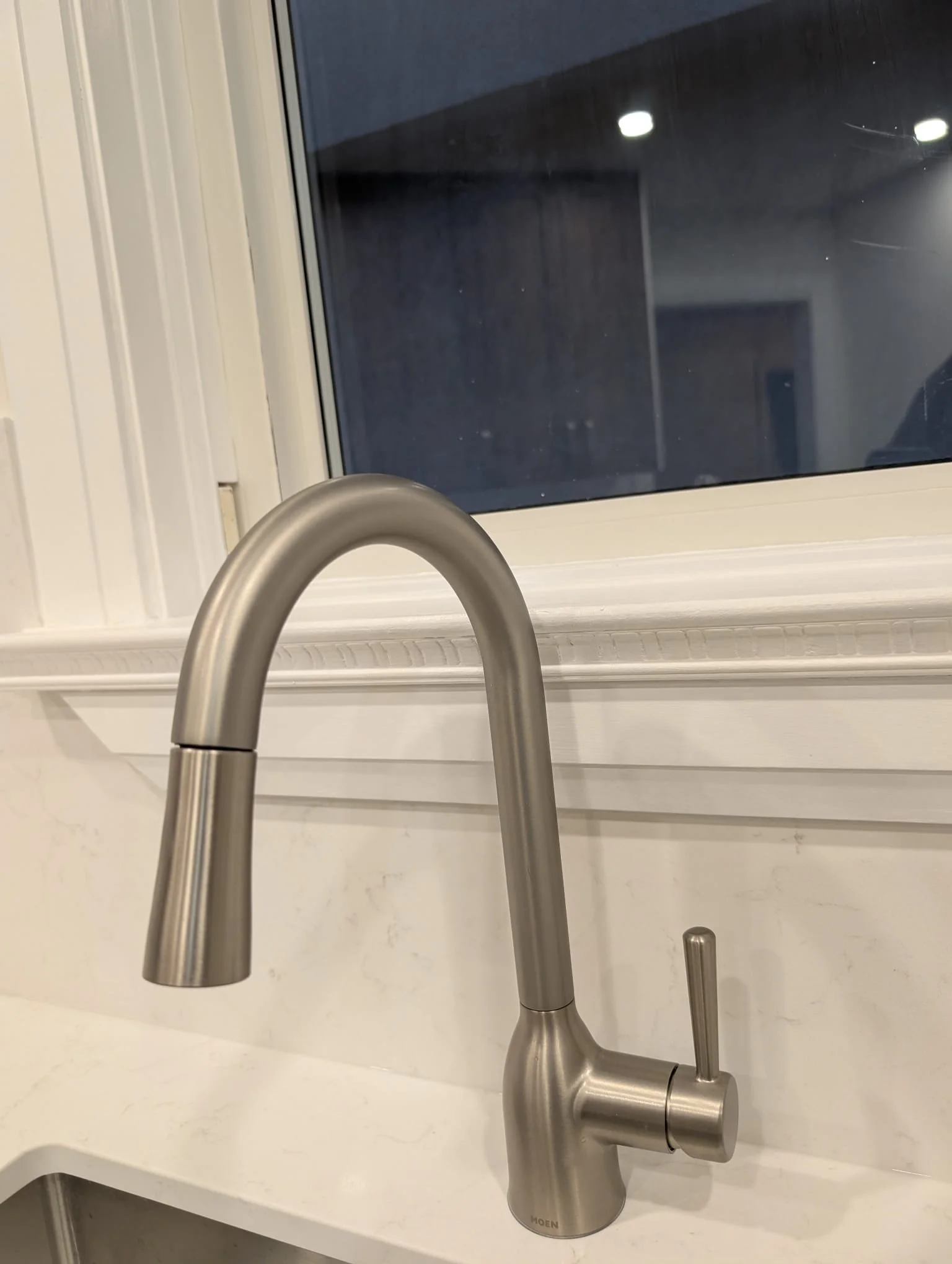 Modern stainless steel kitchen faucet above a white countertop and a window in the background.