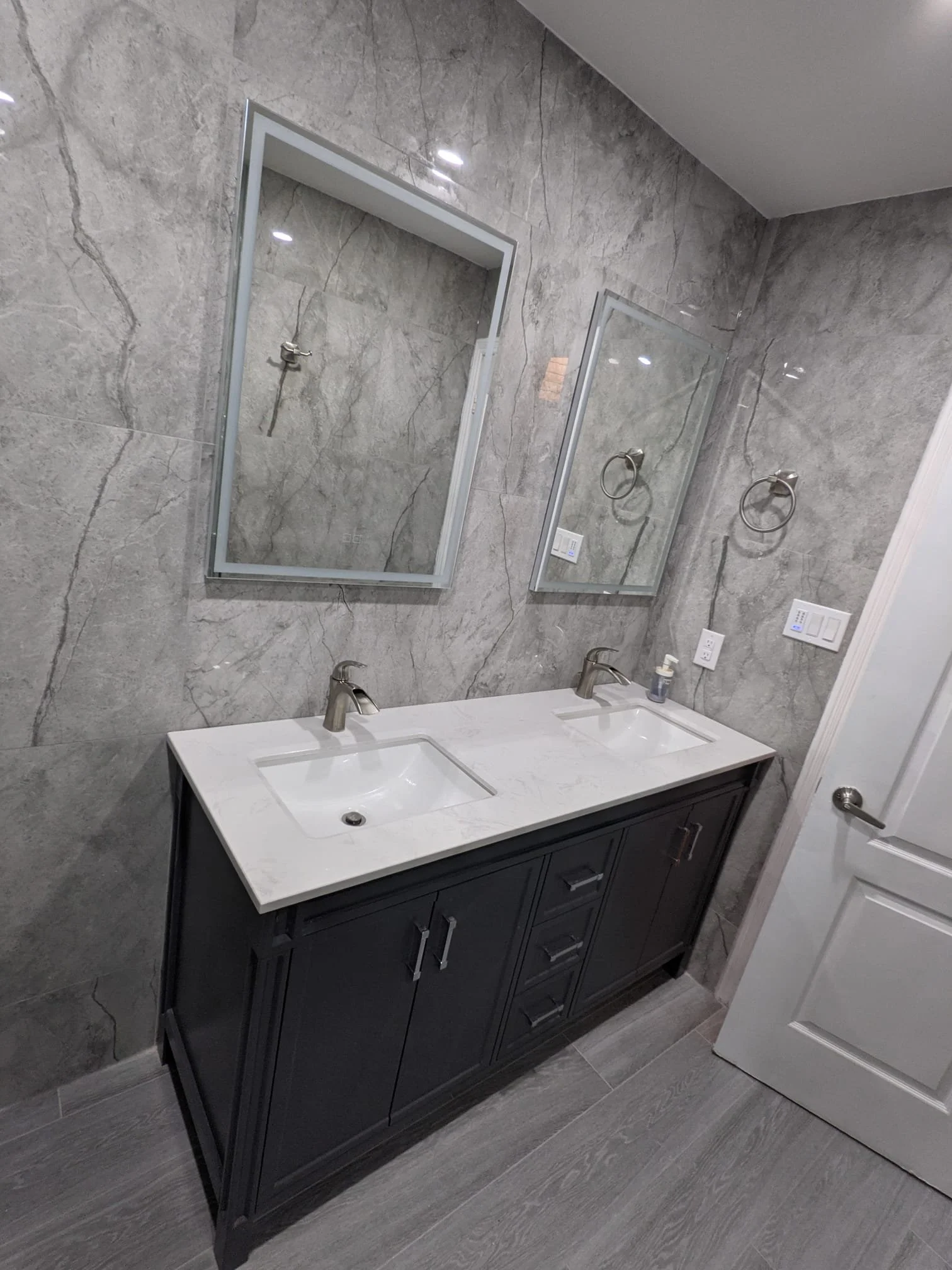 Bathroom with double vanity, gray marble walls, two mirrors, two sinks, and gray wood floor.