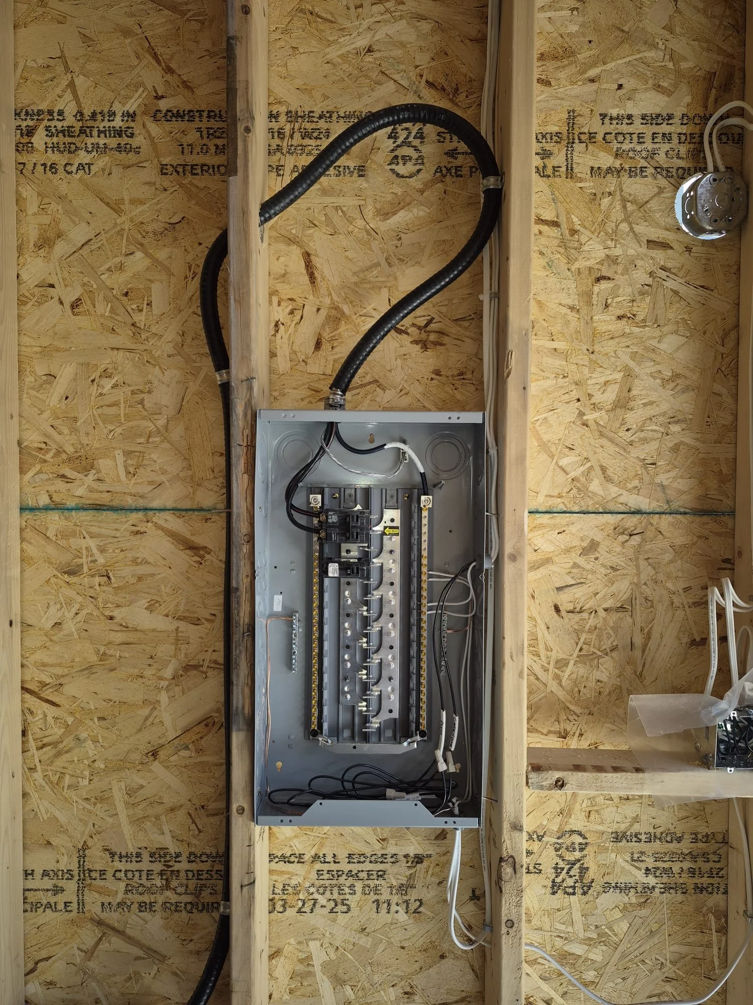 An electrical wiring panel installed in a wall with unfinished wood framing. The panel contains wires and circuit breakers, with a large black conduit pipe connected at the top.