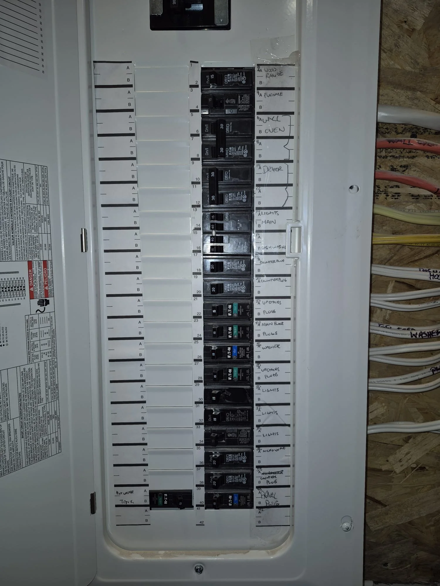 Electrical breaker panel with handwritten labels indicating circuits such as wood, furnace, wall oven, dryer, lights, main, dishwasher, counter, upstairs plugs, main fuse, washer, and panel box.