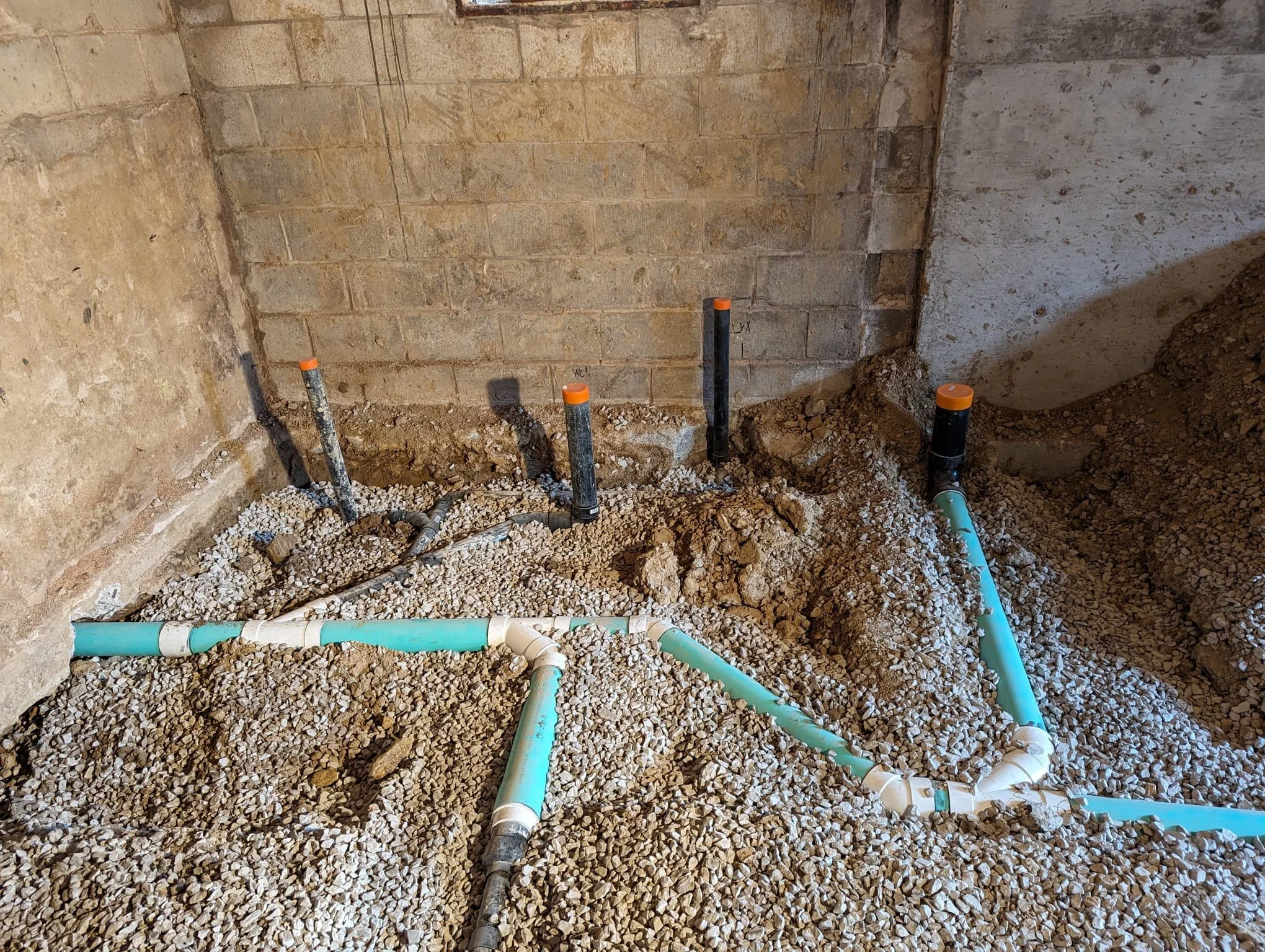 Underground plumbing pipes in a construction site with gravel and dirt, including black and teal pipes with orange caps, against unfinished brick and concrete walls.