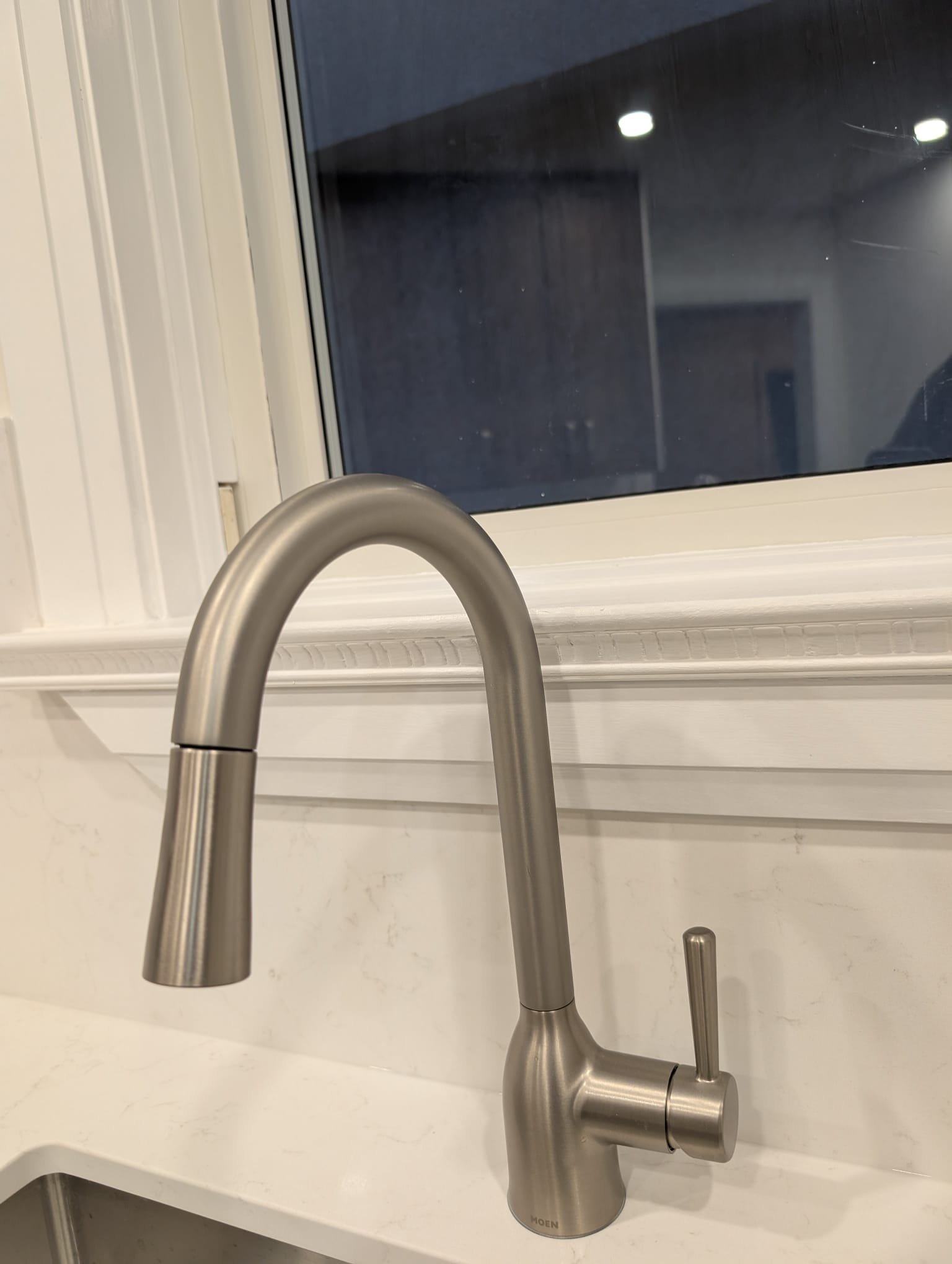 Modern stainless steel kitchen faucet above a white marble countertop near a window.