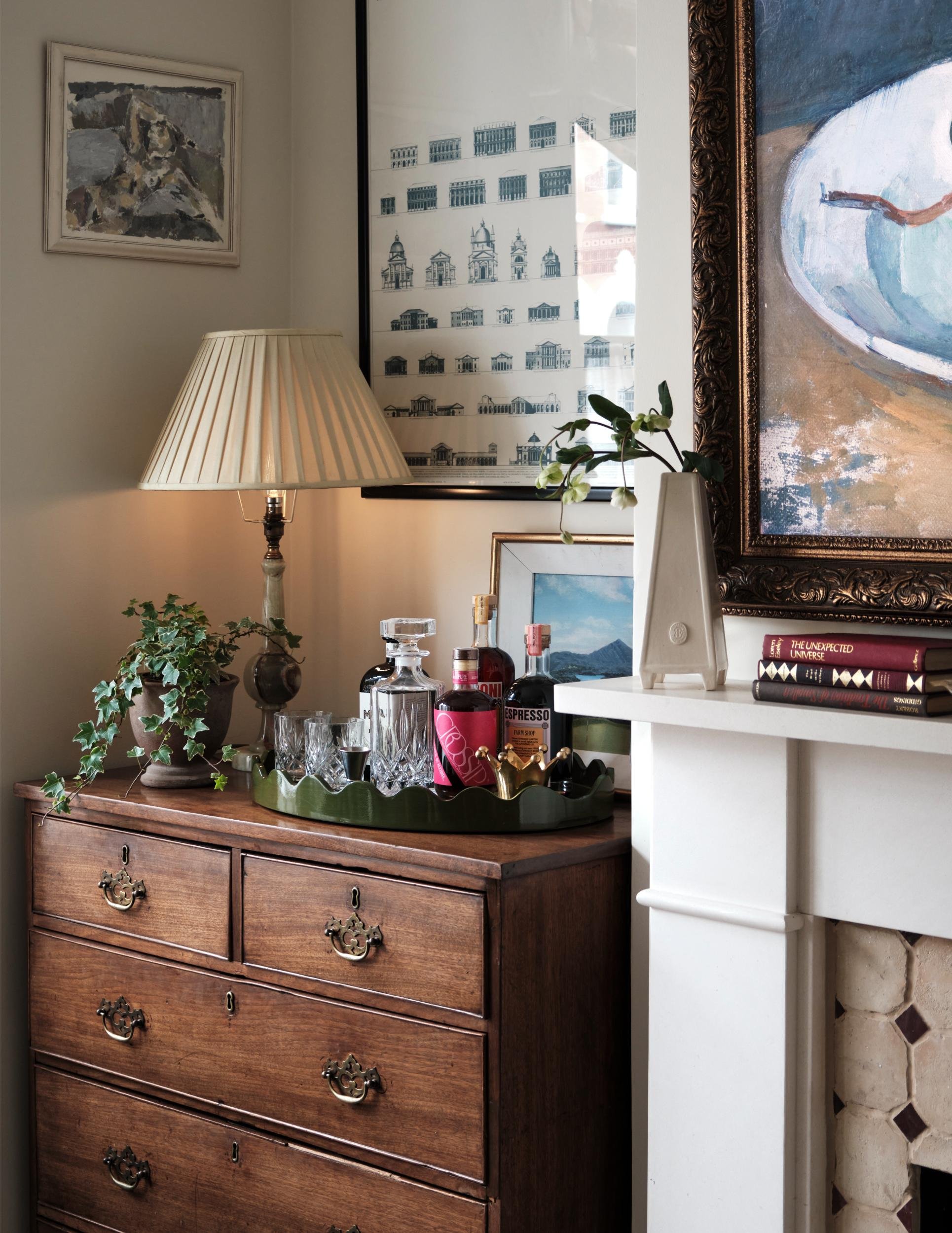A wooden dresser with a green tray holding bottles of liquor and glasses, a plant, a lamp, and framed artwork on the wall.