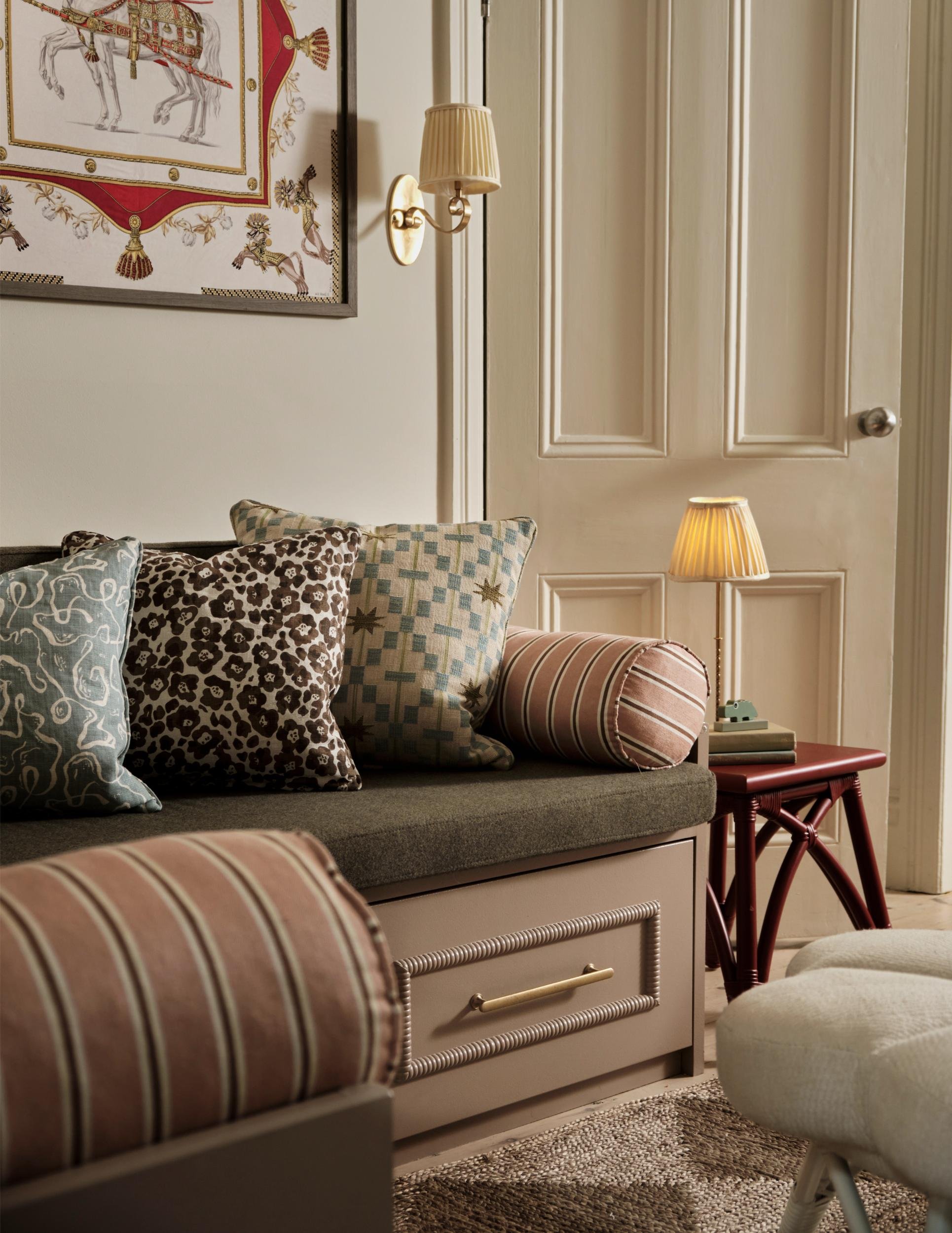Living room with a built-in window seat, decorated with patterned throw pillows, a wall picture, a small side table with a lamp, books, and vintage decor, and a beige door.