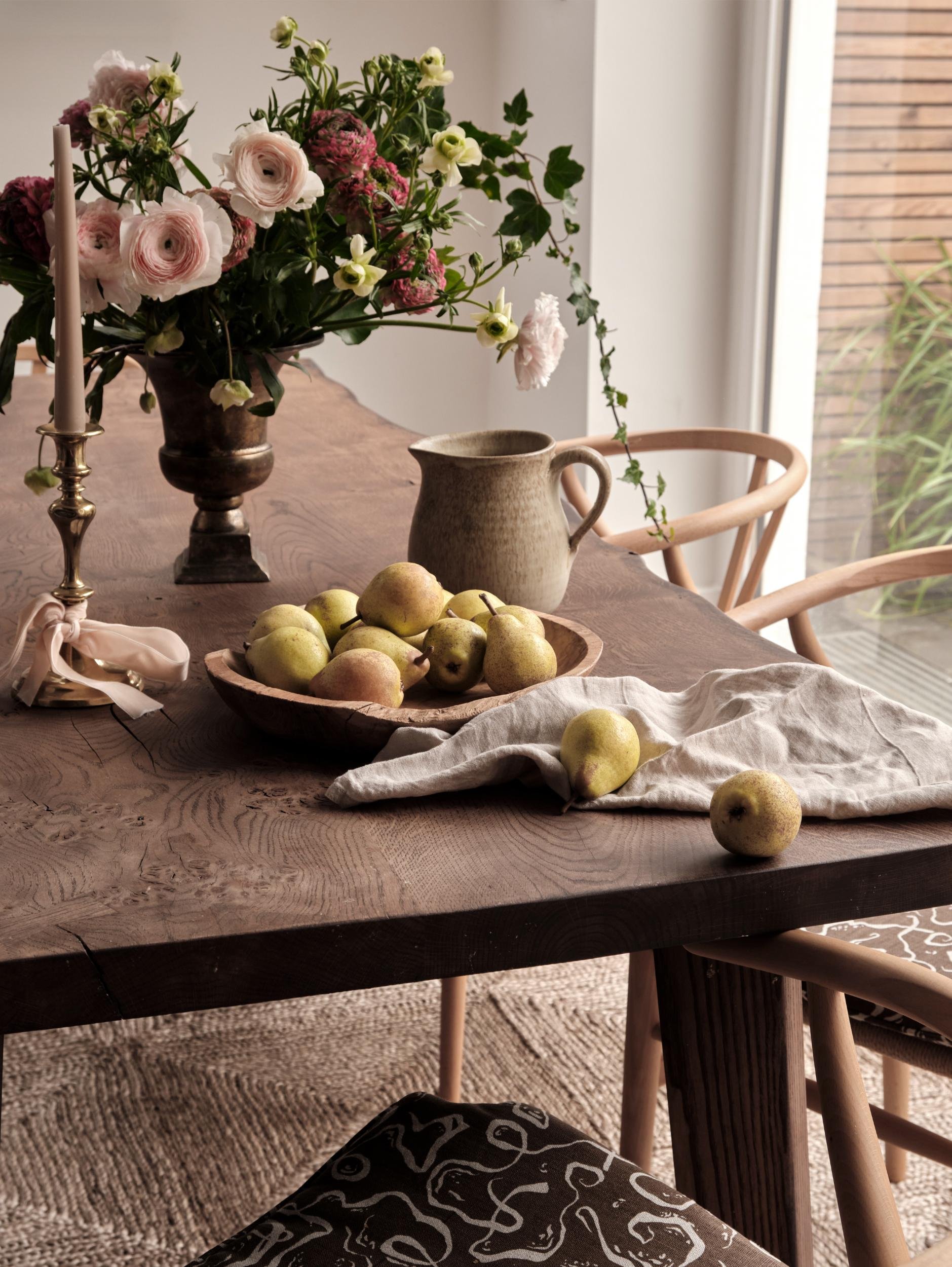 A wooden dining table with a vase of pink and white flowers, a bowl of pears, a ceramic pitcher, and a white cloth. A candlestick with a pink candle is also on the table, near a window with wooden blinds.