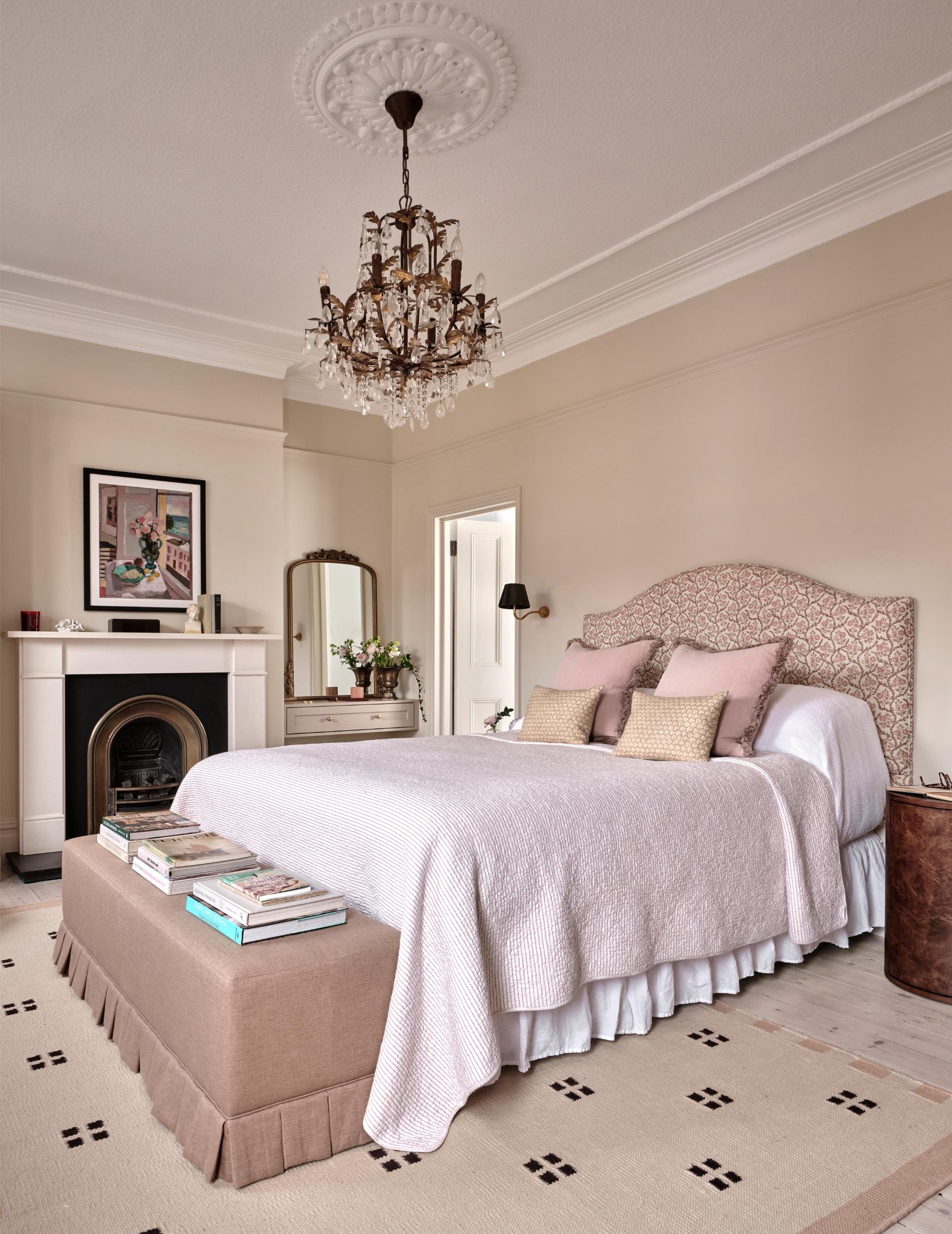 A cozy bedroom with a chandelier, a bed with pink and beige pillows, a beige ottoman topped with books, a fireplace, and a mirror with flowers on a side table.