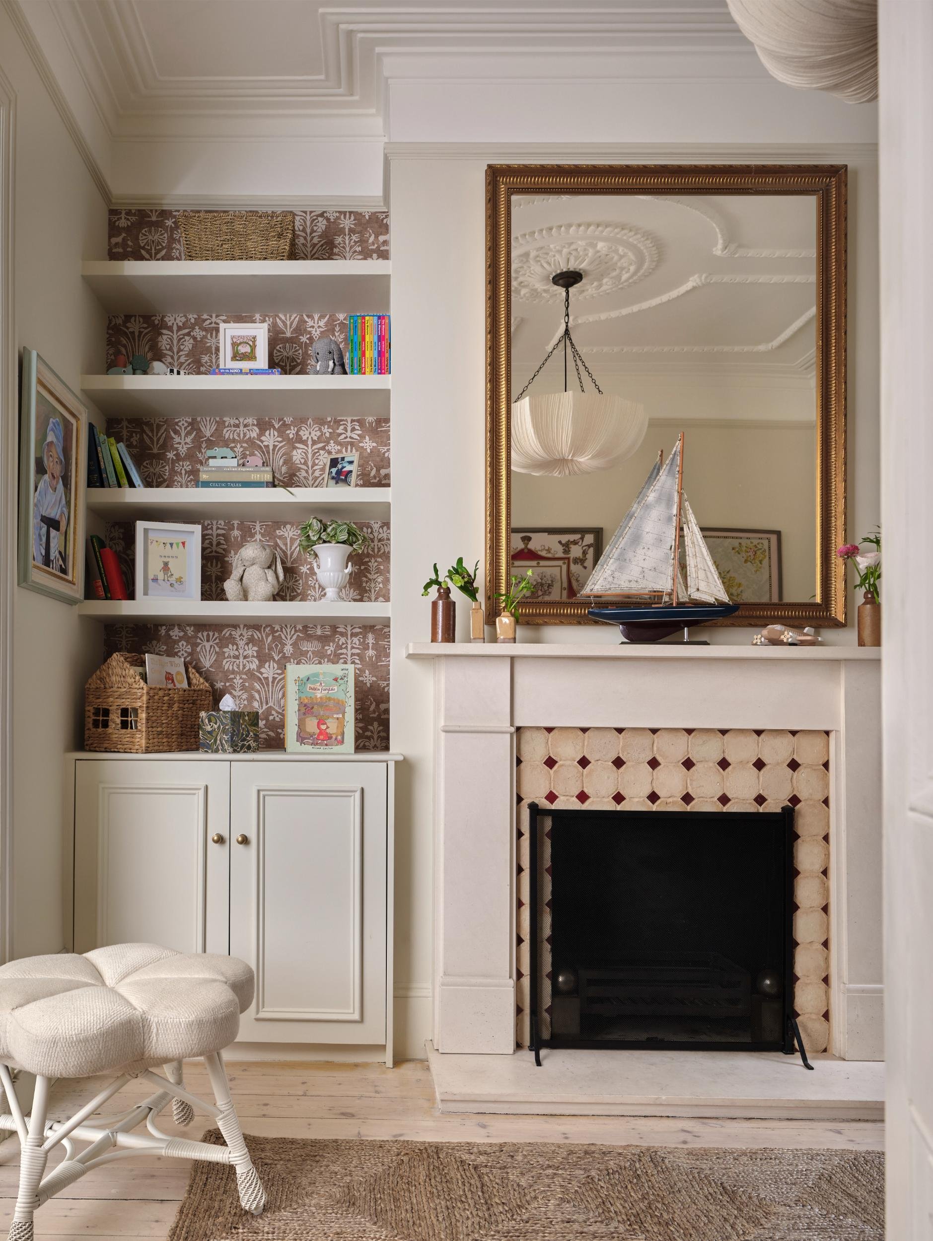 A cozy living room corner with a fireplace, large mirror above it, nautical model boat on mantel, built-in bookshelf with toys and books on the left, tufted white stool in front, and a ceiling light fixture reflected in the mirror.