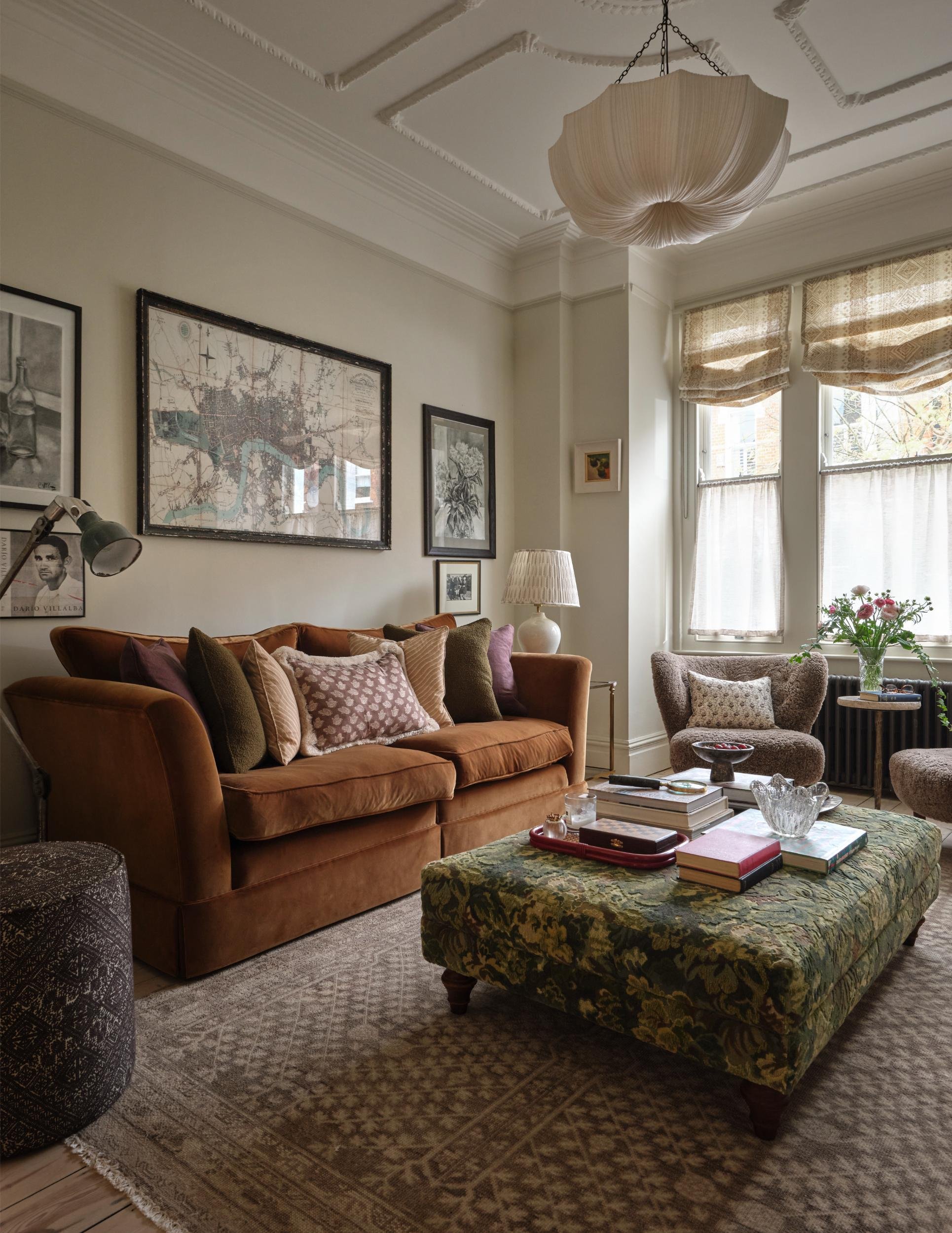 A cozy living room with a vintage look, featuring a brown velvet sofa with multiple pillows, a patterned ottoman with books and decor on top, a plush armchair by a window with floral curtains, a side table with a flower vase, wall art, and a unique f
