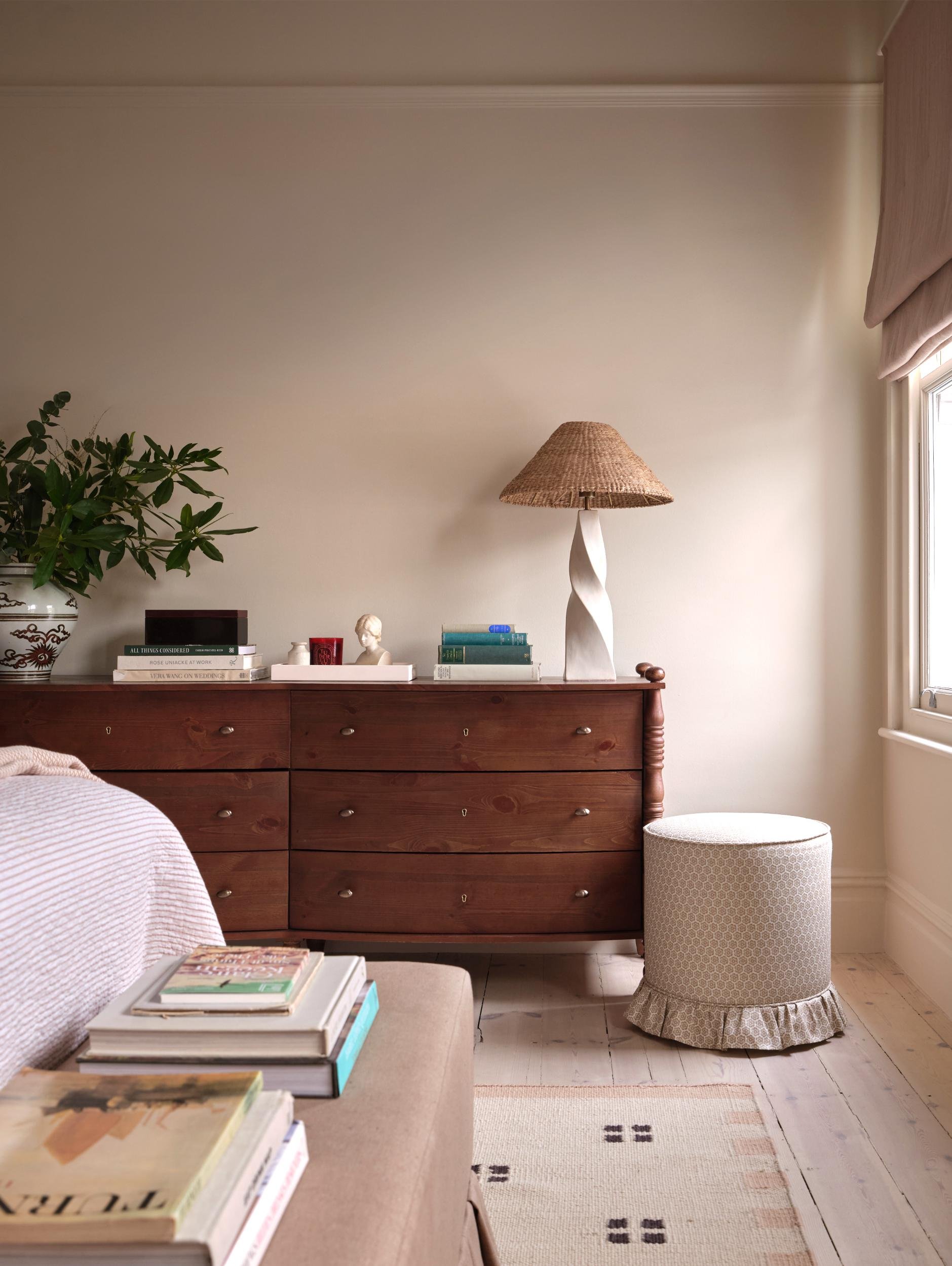 A cozy living room corner with a wooden dresser topped with books, decorative items, a table lamp with a twisted white base and a woven shade, and a potted plant. A round, upholstered storage ottoman with a ruffled skirt sits next to a window with be