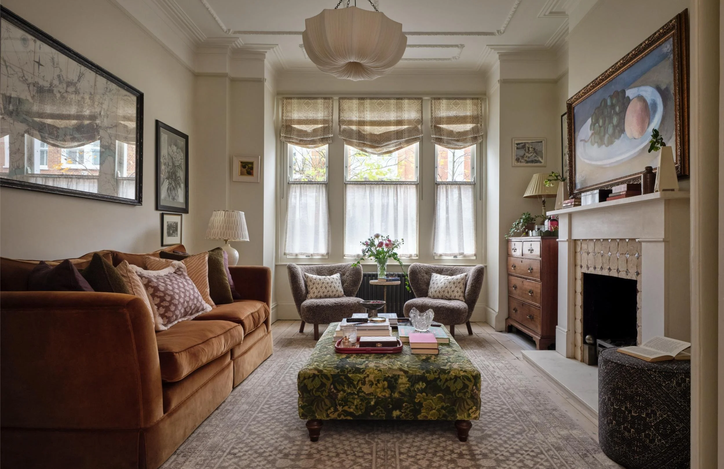 Cozy living room with a brown velvet sofa, two armchairs near a large window, a floral ottoman, and a fireplace with artwork on the mantle.