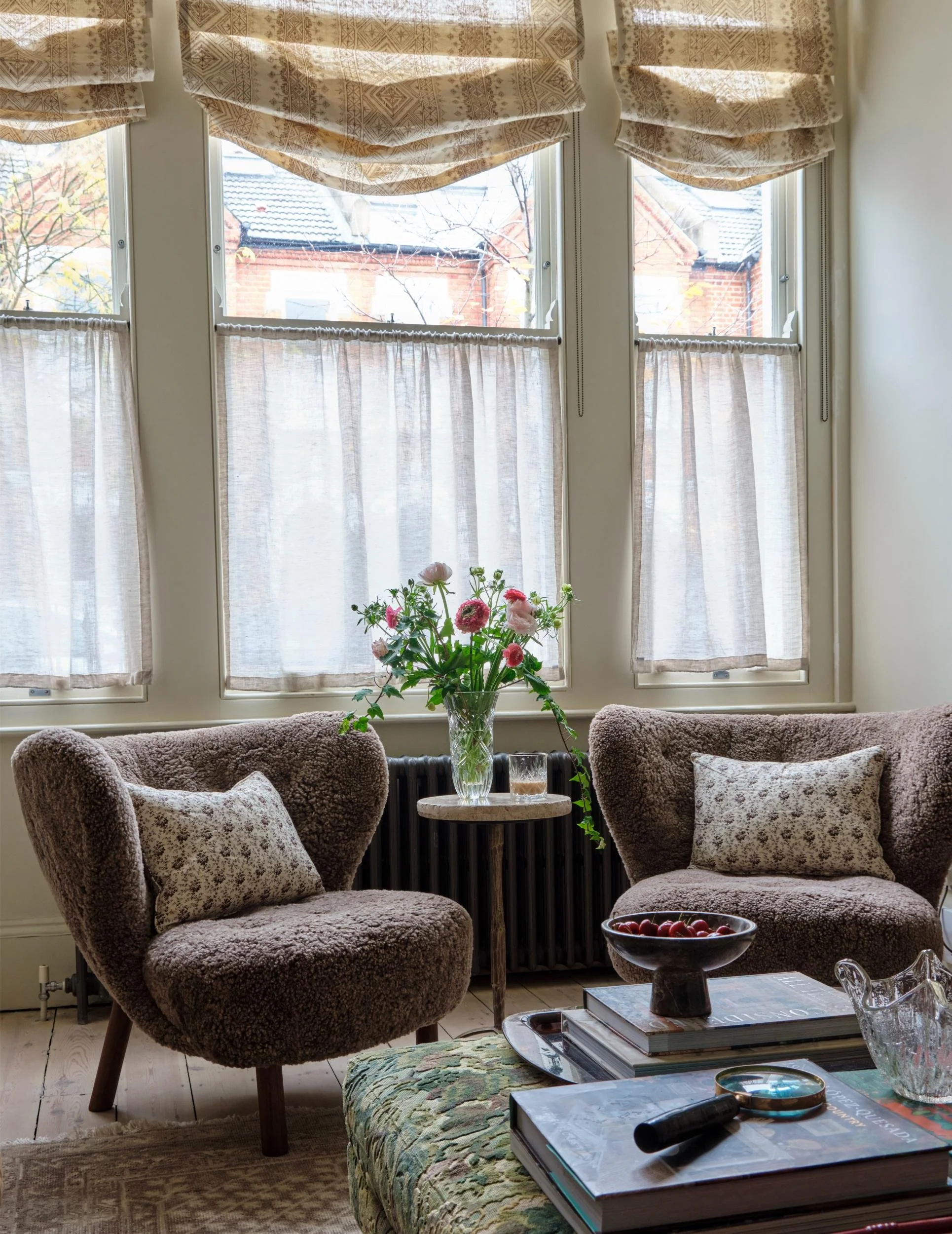 Cozy living room with two plush brown armchairs with patterned pillows, a small round wooden table with a vase of pink flowers, a bowl of cherries, and a stack of magazines on an upholstered ottoman near a large window with beige curtains and a view of trees and houses outside.