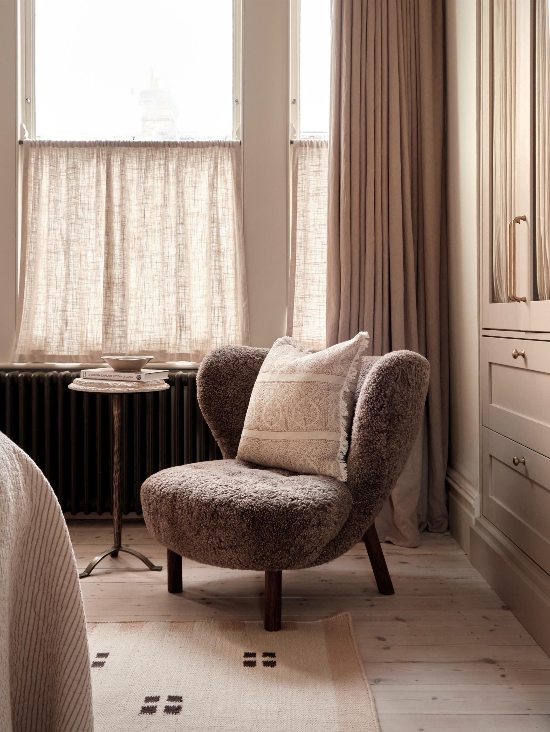 A cozy corner with a plush, brown upholstered armchair with a matching pillow, next to a small side table with a book and a decorative bowl, near large windows with cream-colored curtains and a beige room rug.
