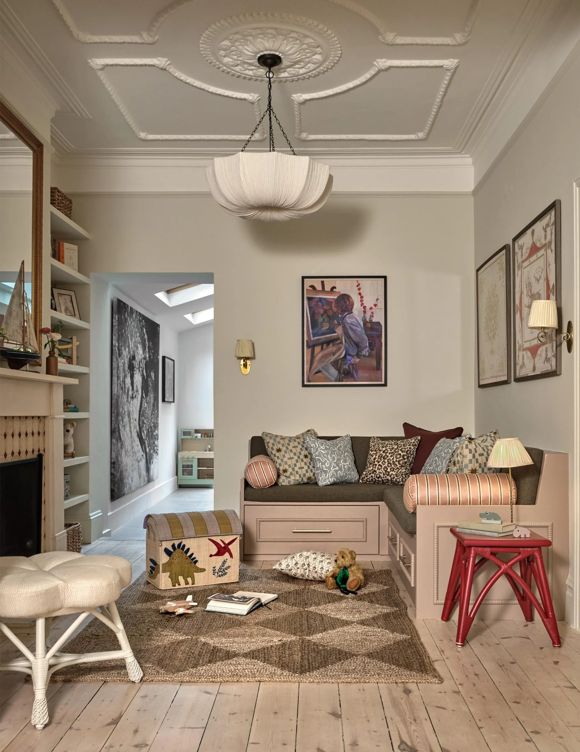 A cozy living room with patterned pillows on a built-in bench, framed artwork on the wall, a hanging ceiling light, and toys on a plush rug, with a doorway leading to a play area featuring a small kitchen set.