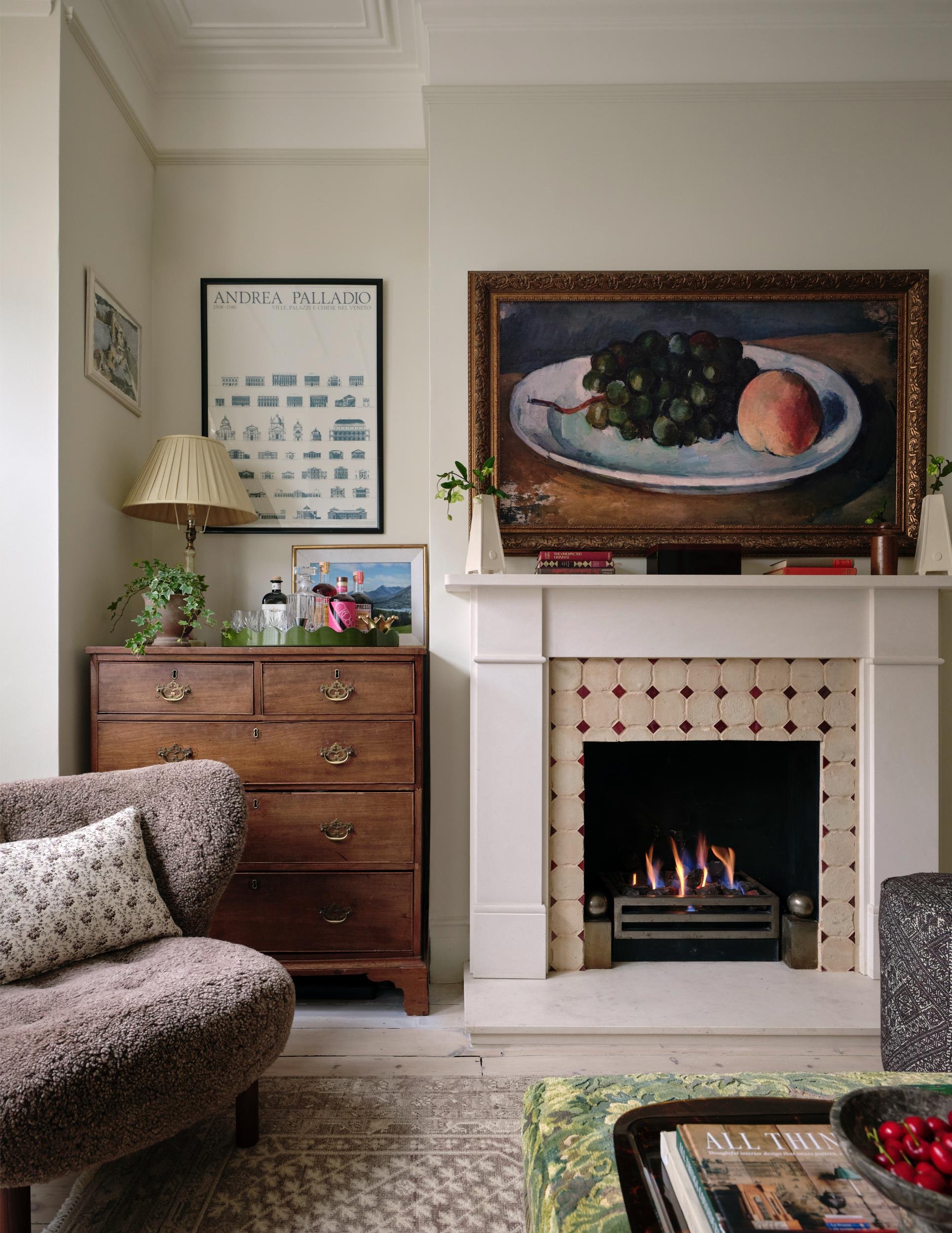 Living room with a fireplace, a framed still life painting of grapes and a peach above it, a wooden dresser with plants and bottles, a beige armchair with a pillow, and a patterned ottoman with books and a bowl of cherries.