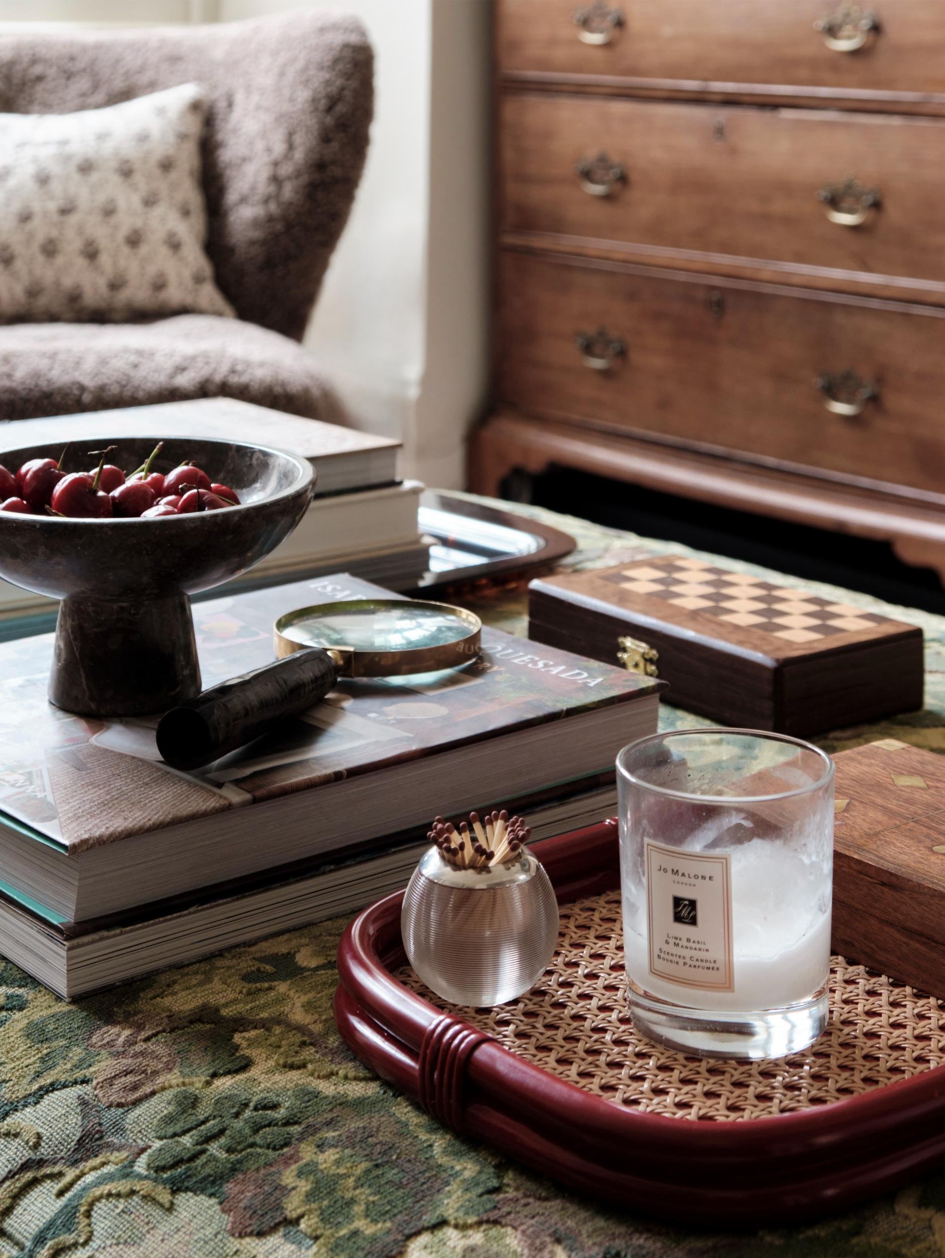 A cluttered coffee table with a large book, a bowl of cherries, a jar of decorative matches, a candle, a chessboard, and a glass with a candle inside, situated in a room with a wooden dresser and a beige upholstered sofa.