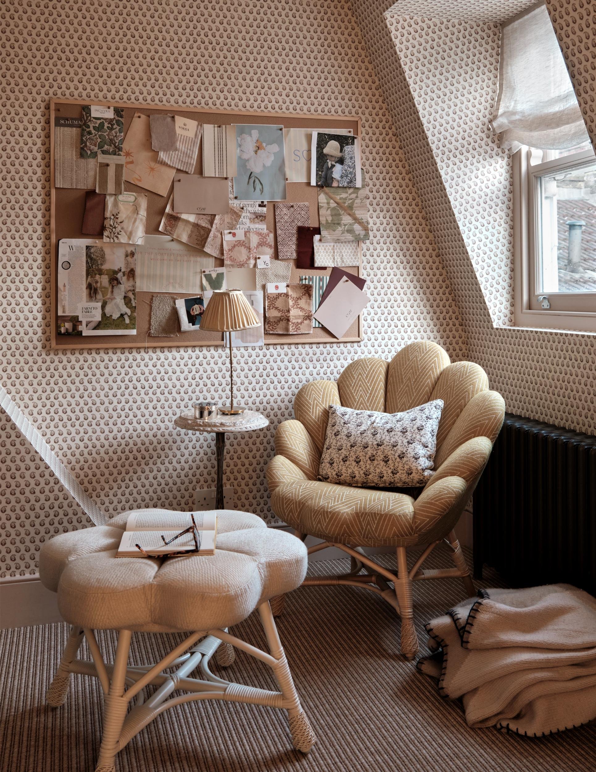 A cozy room with patterned wallpaper, a bulletin board filled with fabric swatches and photos, a small lamp on a round side table, a cream-colored tufted stool with an open book and glasses on top, a mustard yellow armchair with a floral pillow, a bl