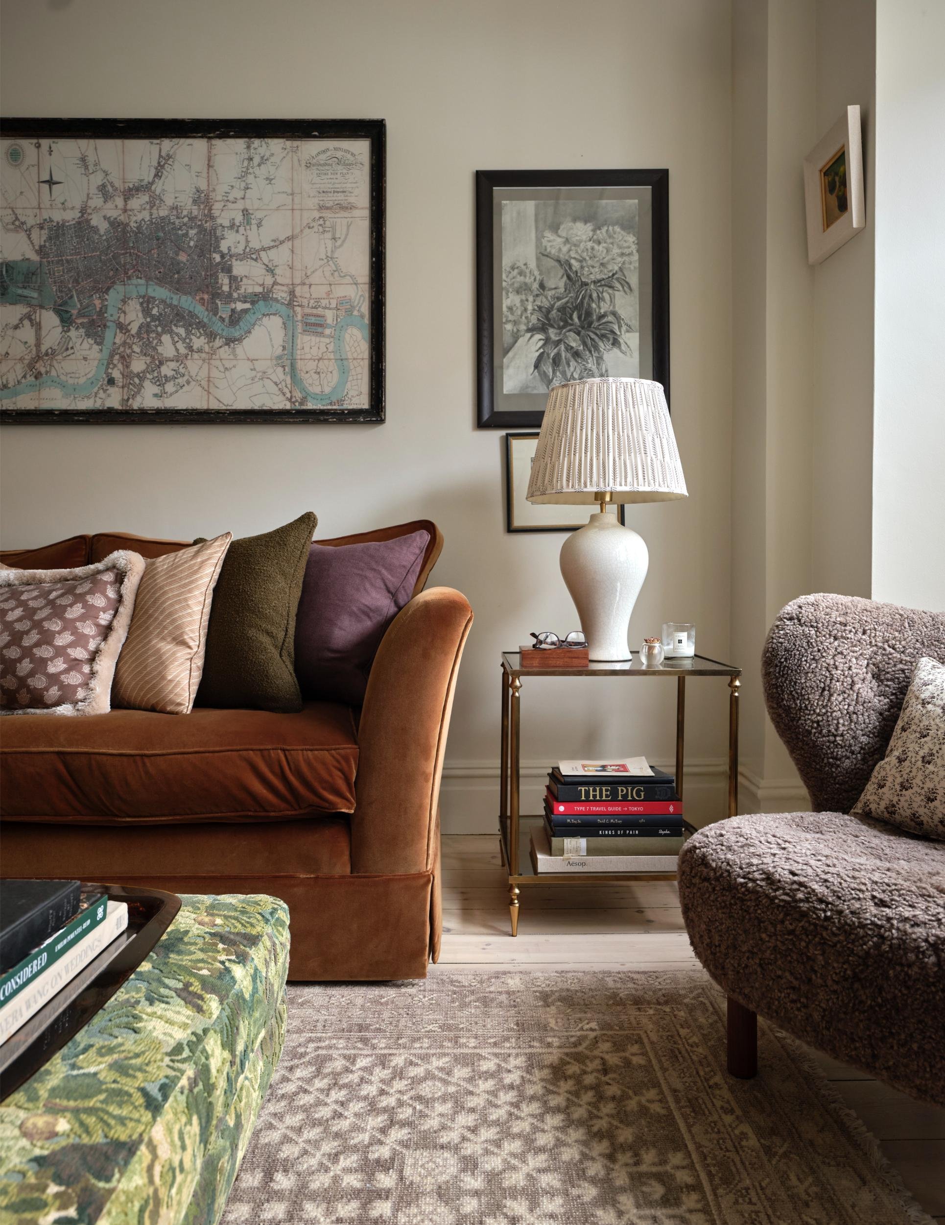 Interior of a living room with a brown velvet sofa, patterned cushions, a side table with a white ceramic lamp, glasses, and books, and framed artwork on the wall.