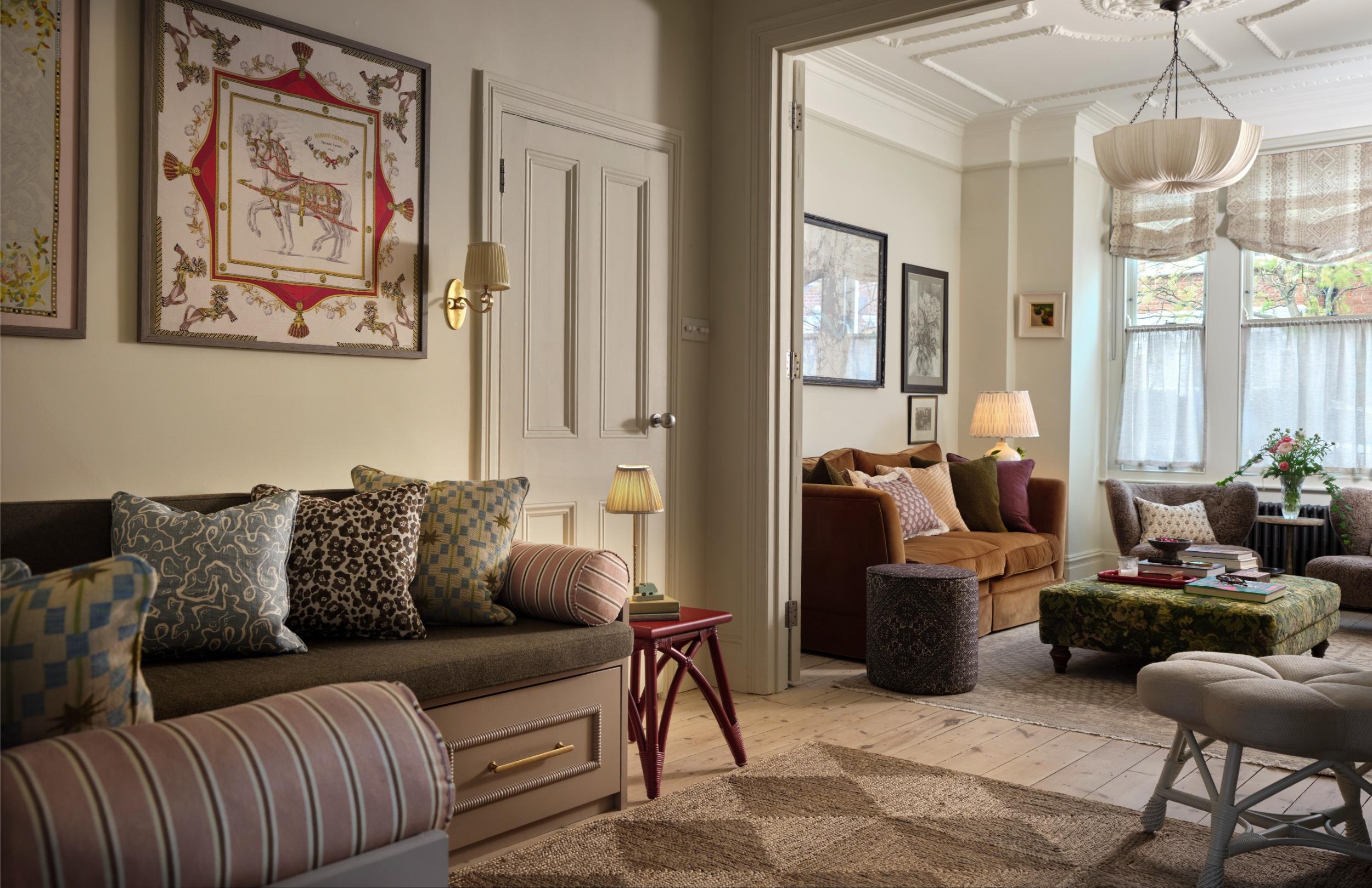 Living room with cozy seating, colorful cushions, side tables with lamps, artwork on the walls, and large windows with curtains.