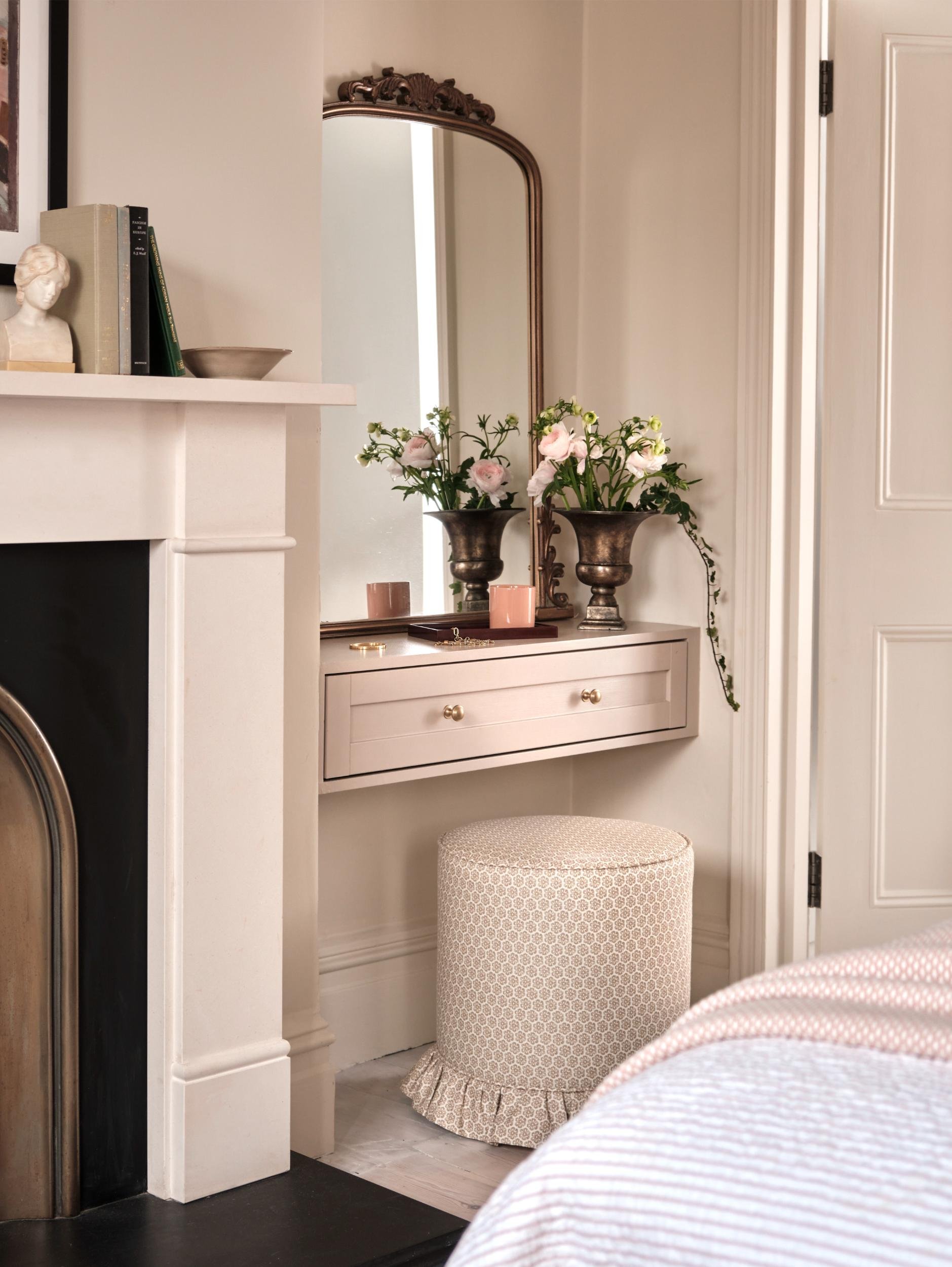 A cozy bedroom corner with a white vanity table, a fireplace mantel with books and a bust sculpture, a full-length mirror, pink flowers in two dark brass vases, a pink candle, a small box with jewelry, and a patterned round ottoman with a ruffled ski