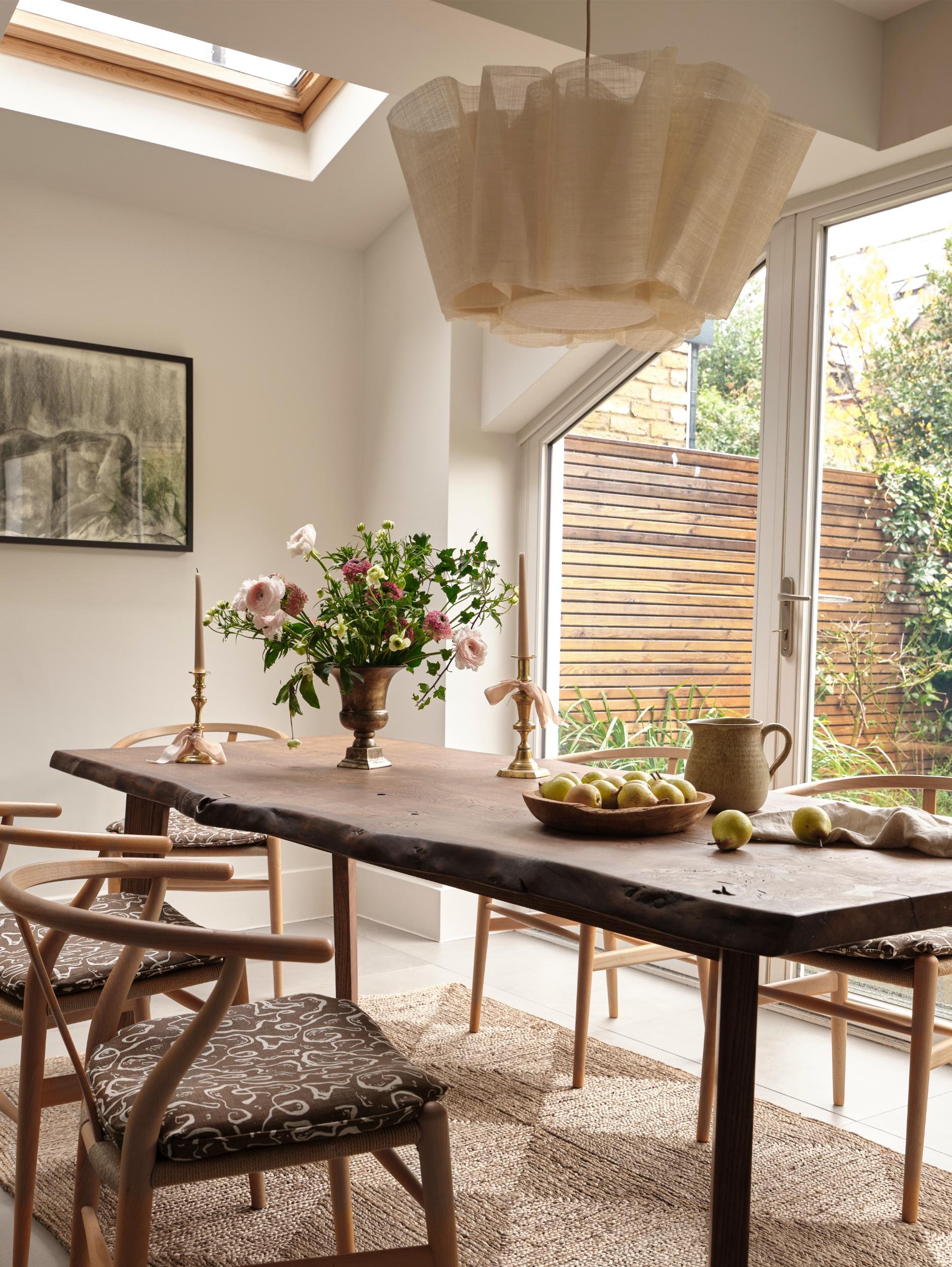A dining room with a rustic wooden table, six chairs with patterned cushions, a vase of pink and white flowers, a bowl of green apples, and two brass candlesticks with white candles. Large windows and a skylight bring in natural light, and a framed b