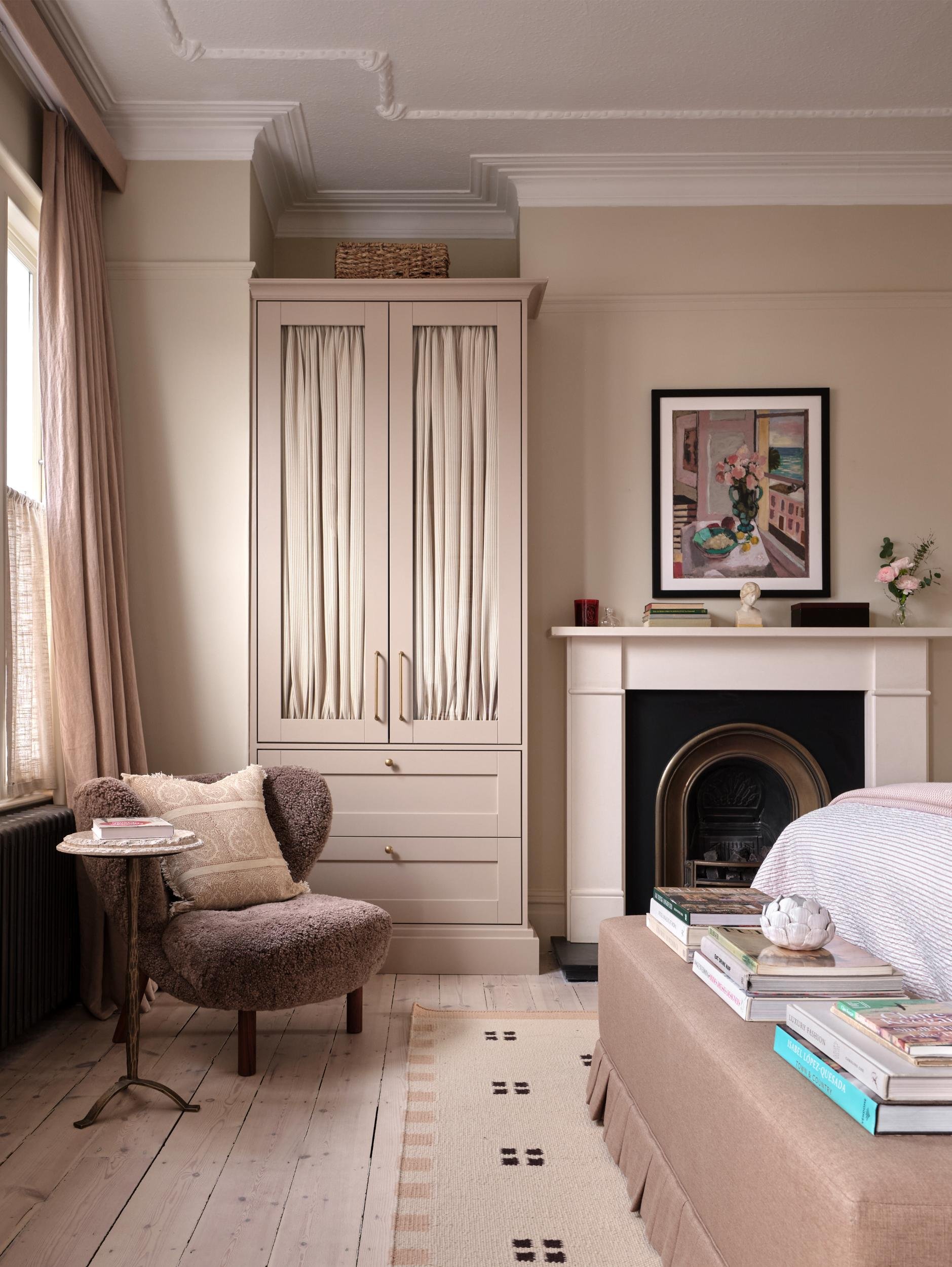 A cozy bedroom corner with a brown armchair, a small side table with books, a white cabinet with curtains, a fireplace with a picture above, and a beige bed with stacked books and a decorative bowl.