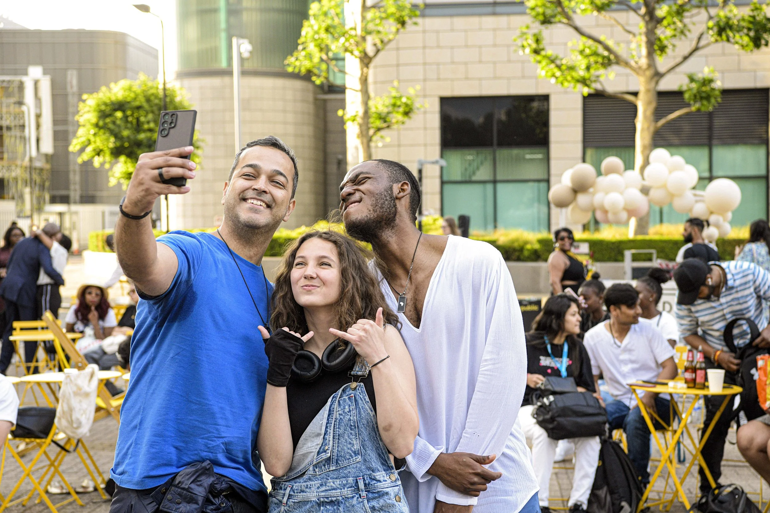 Three people taking a selfie outdoors at a lively event with many people, trees, and modern buildings in the background.