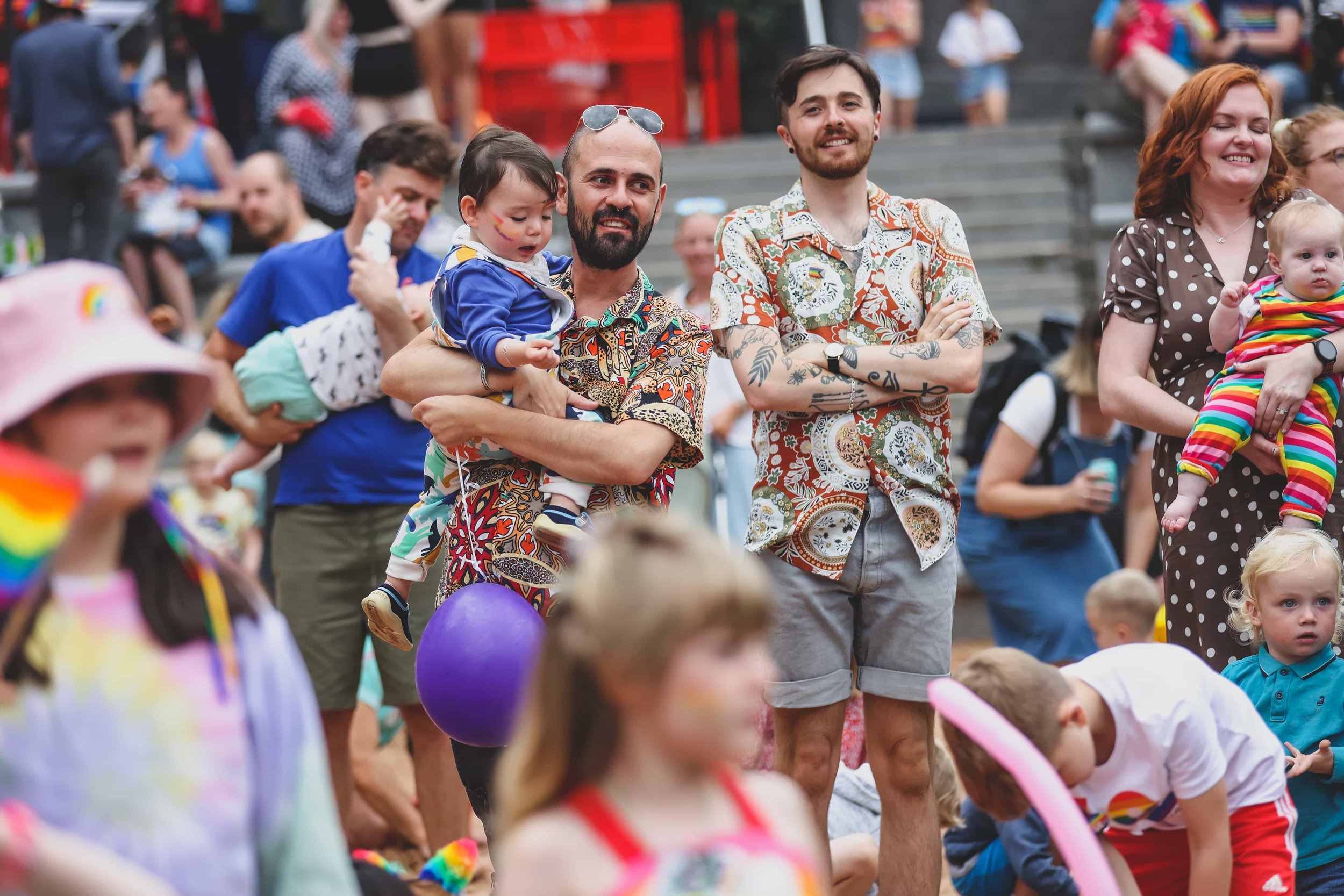 People gathered outdoors at a pride event, some holding children, with rainbow-themed accessories and decorations.