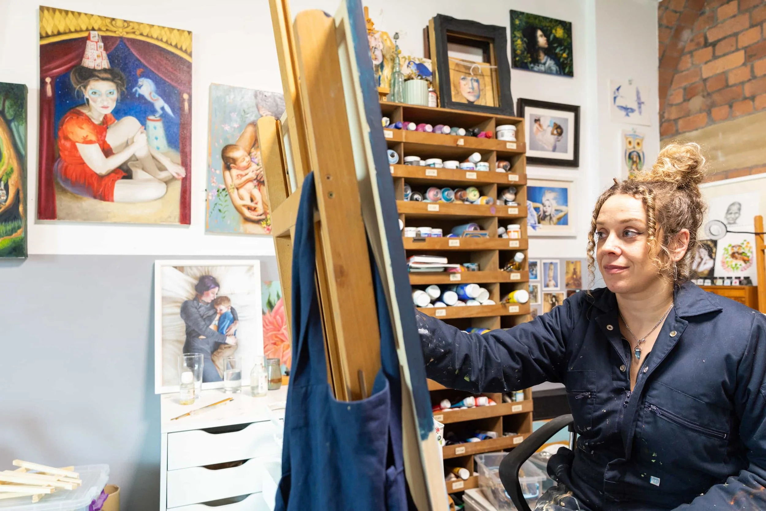 An artist with curly hair in a bun painting in a colorful art studio filled with paintings and art supplies.