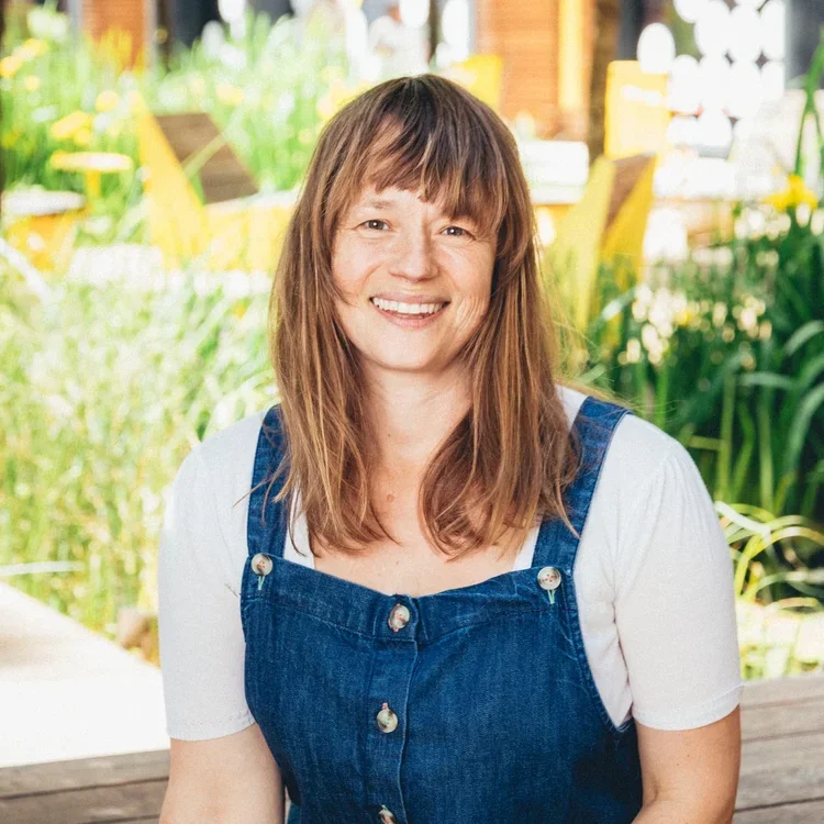Smiling woman with shoulder-length brown hair and bangs, wearing a white shirt and denim overalls, sitting outdoors in a garden or park setting with greenery and yellow structures in the background.