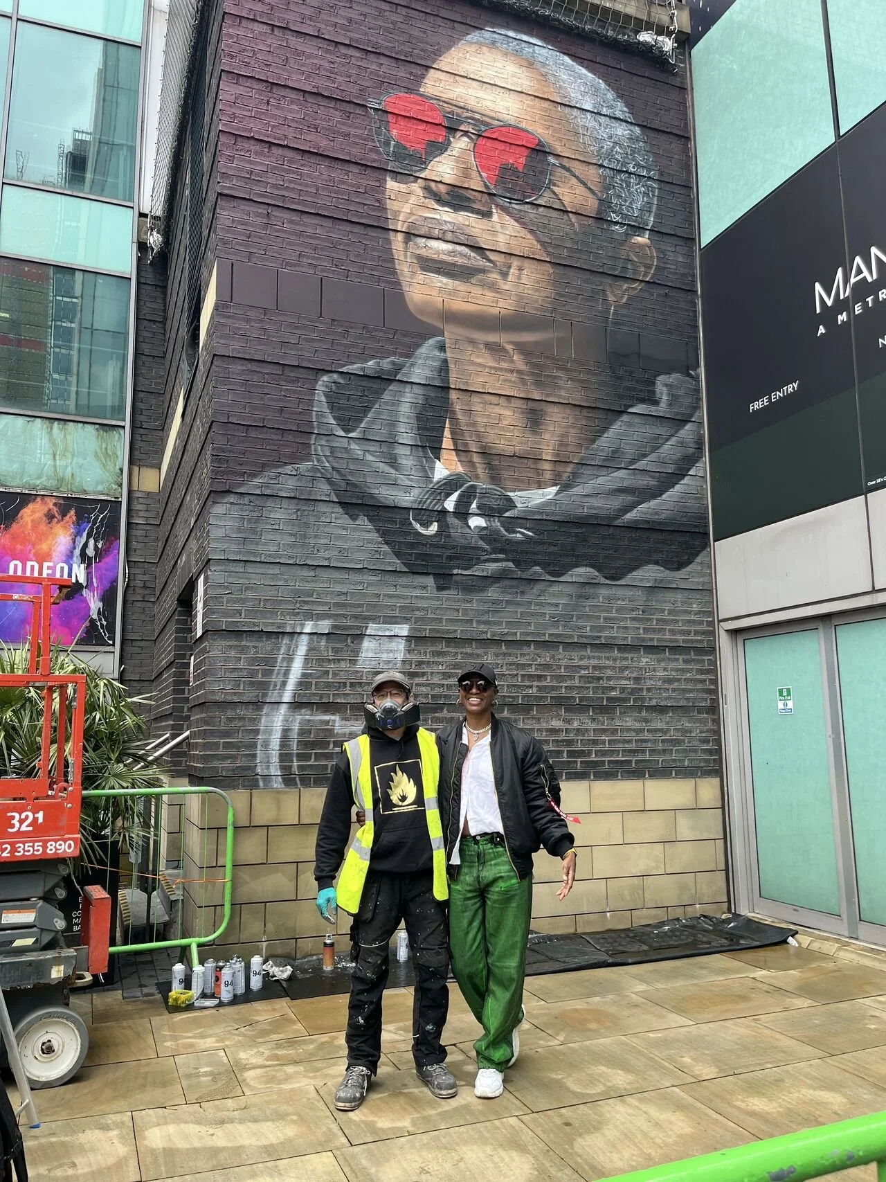 Two people standing in front of a large mural of a woman wearing sunglasses on a brick wall.