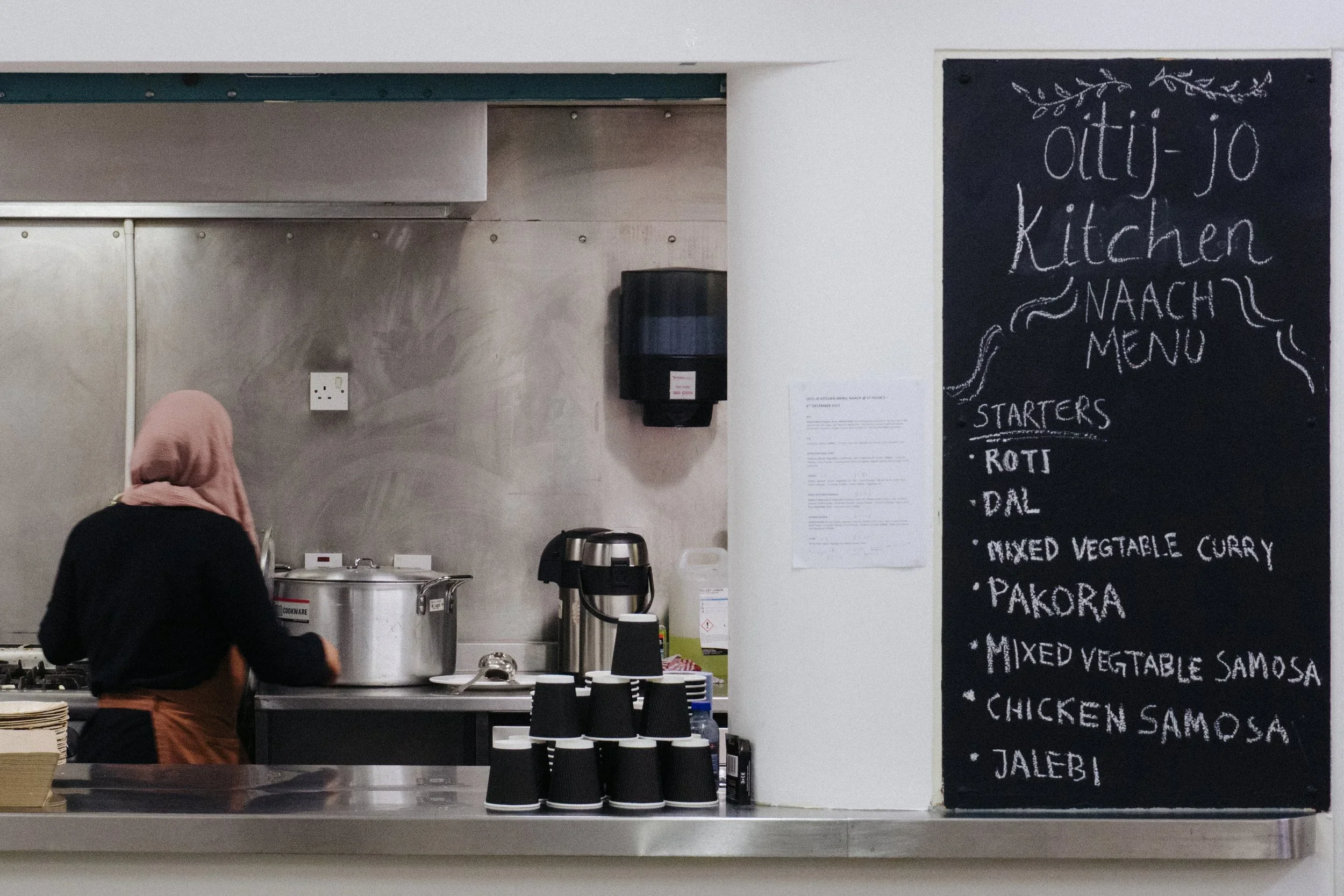 A woman wearing a headscarf and apron cooking in a kitchen with a large pot on the stove. To the right, a black chalkboard displays a menu for 'Otti-Jo Kitchen' listing starters such as roti, dal, mixed vegetable curry, pakora, mixed vegetable samosa, chicken samosa, and jalebi.