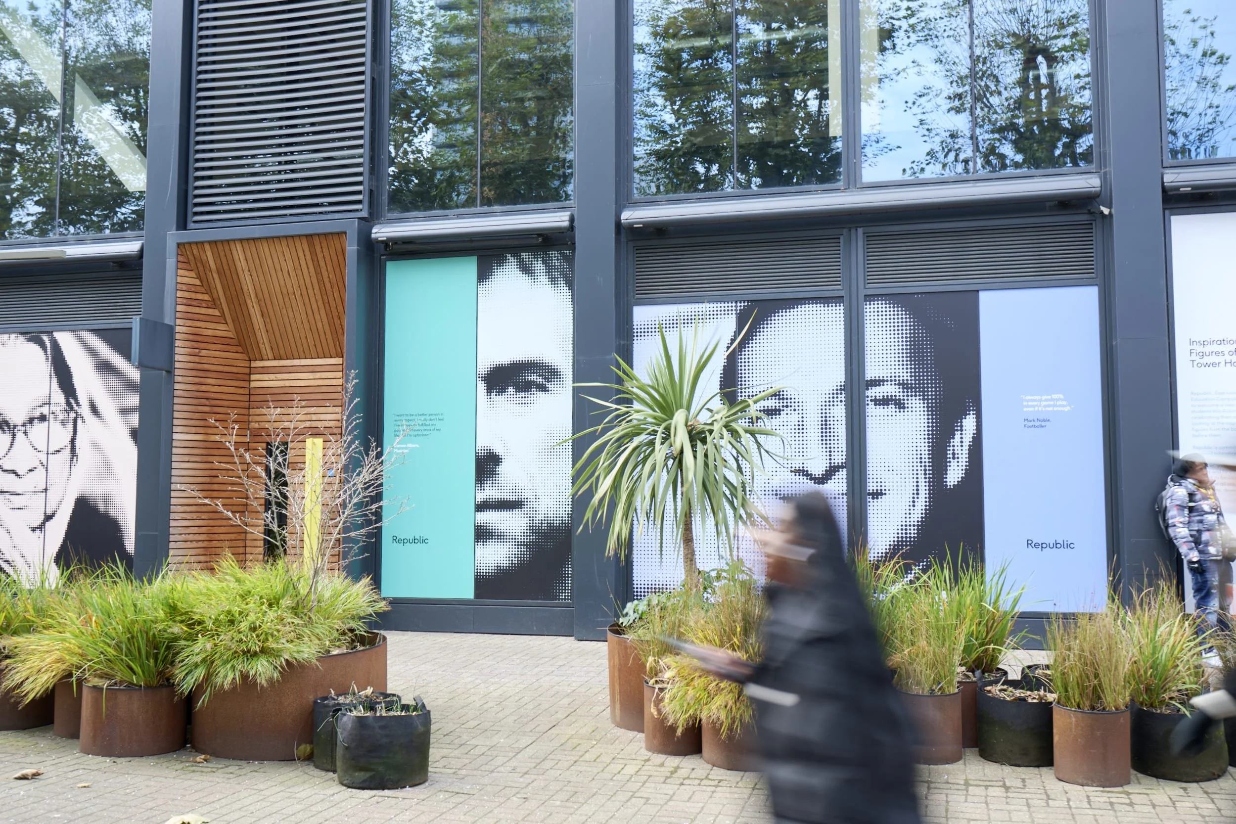 Exterior of a modern building with large graphic portraits of two people on the walls, surrounded by potted plants and blurred pedestrians walking by.