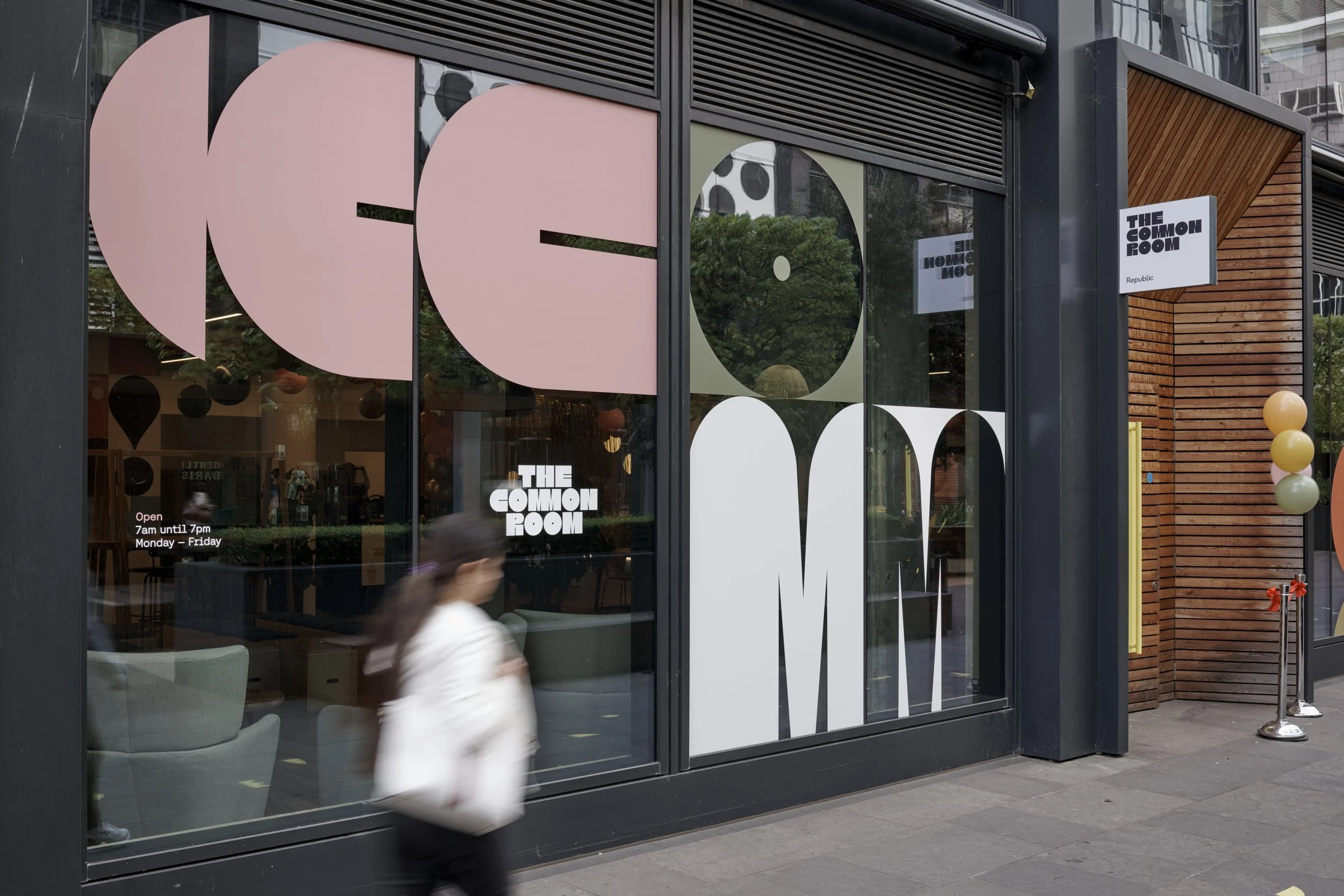 The storefront of a business called The Common Room with large decorative glass window panels and a small sign indicating the operating hours from 7 am to 7 pm, Monday through Friday. A person in a white jacket is walking past the window.