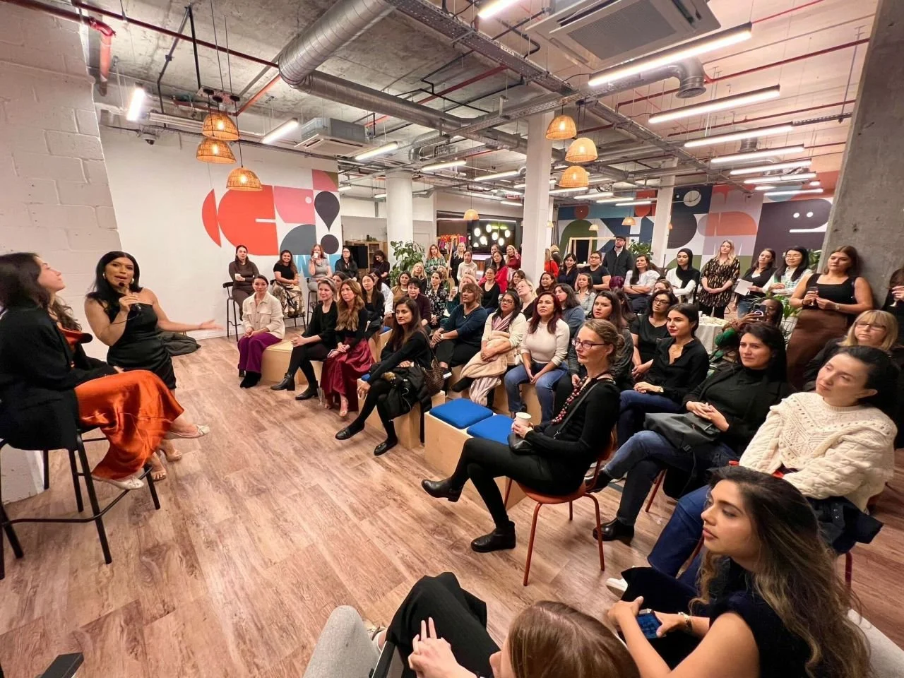 A large group of women attending a discussion or presentation in a modern, decorated indoor space with colorful geometric wall art and warm lighting.