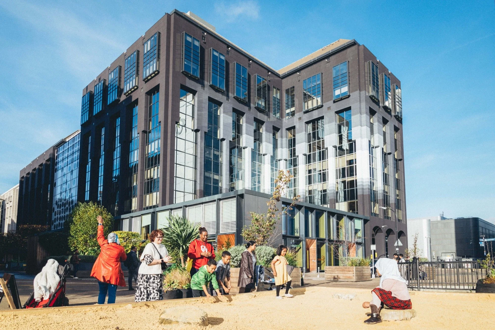 A group of diverse people, including children and adults, are gathered outdoors near a city modern building. Some are standing, some are crouched down, and one person is sitting on a rock, all engaging in activities such as talking, playing, or using their phones. The building in the background is tall with reflective glass windows, and the sky is clear and blue.