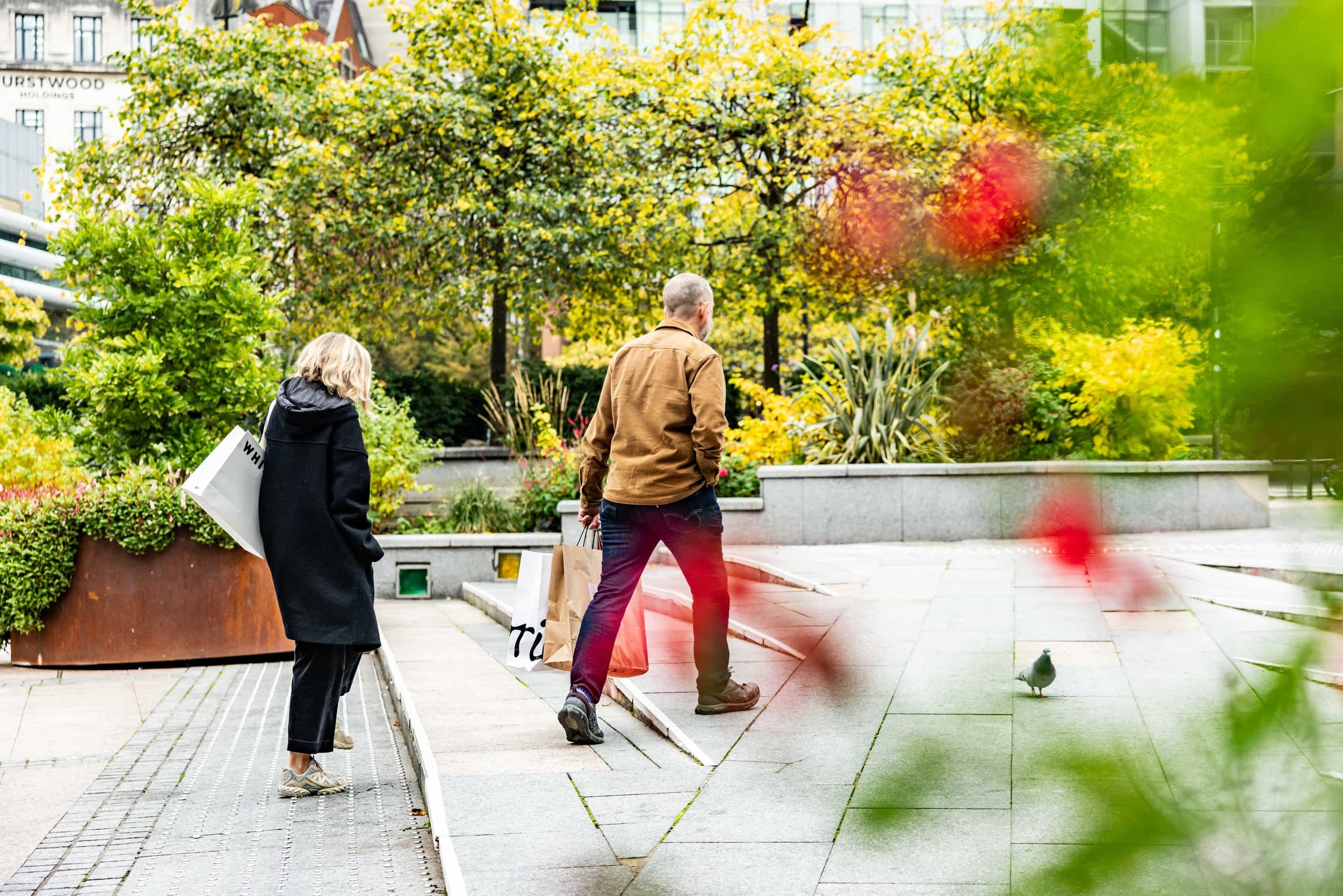 Two people walking on a city sidewalk in a park, surrounded by trees and plants, with a pigeon nearby.