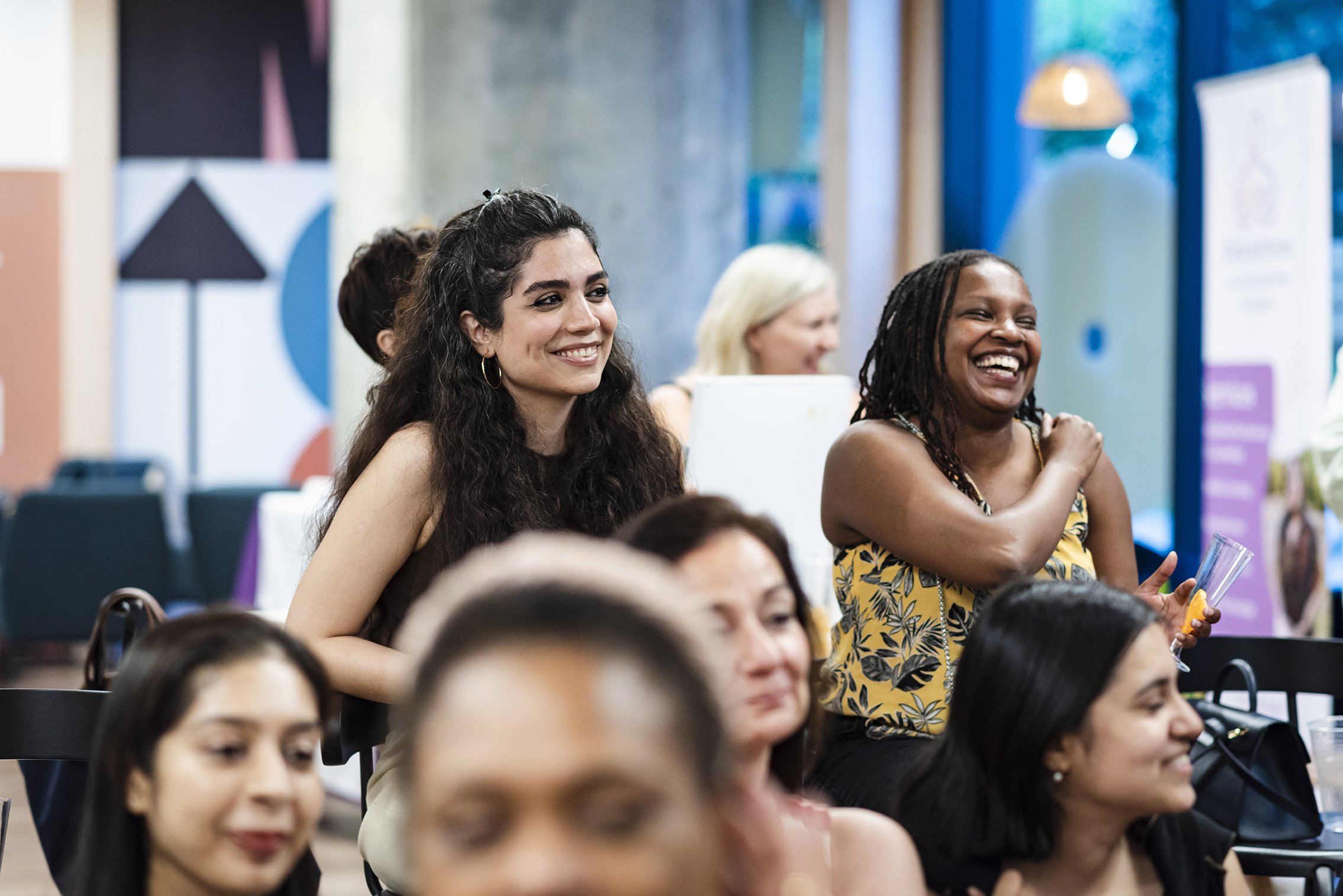 A group of women sitting and standing in a room, smiling and laughing, during an event or gathering.