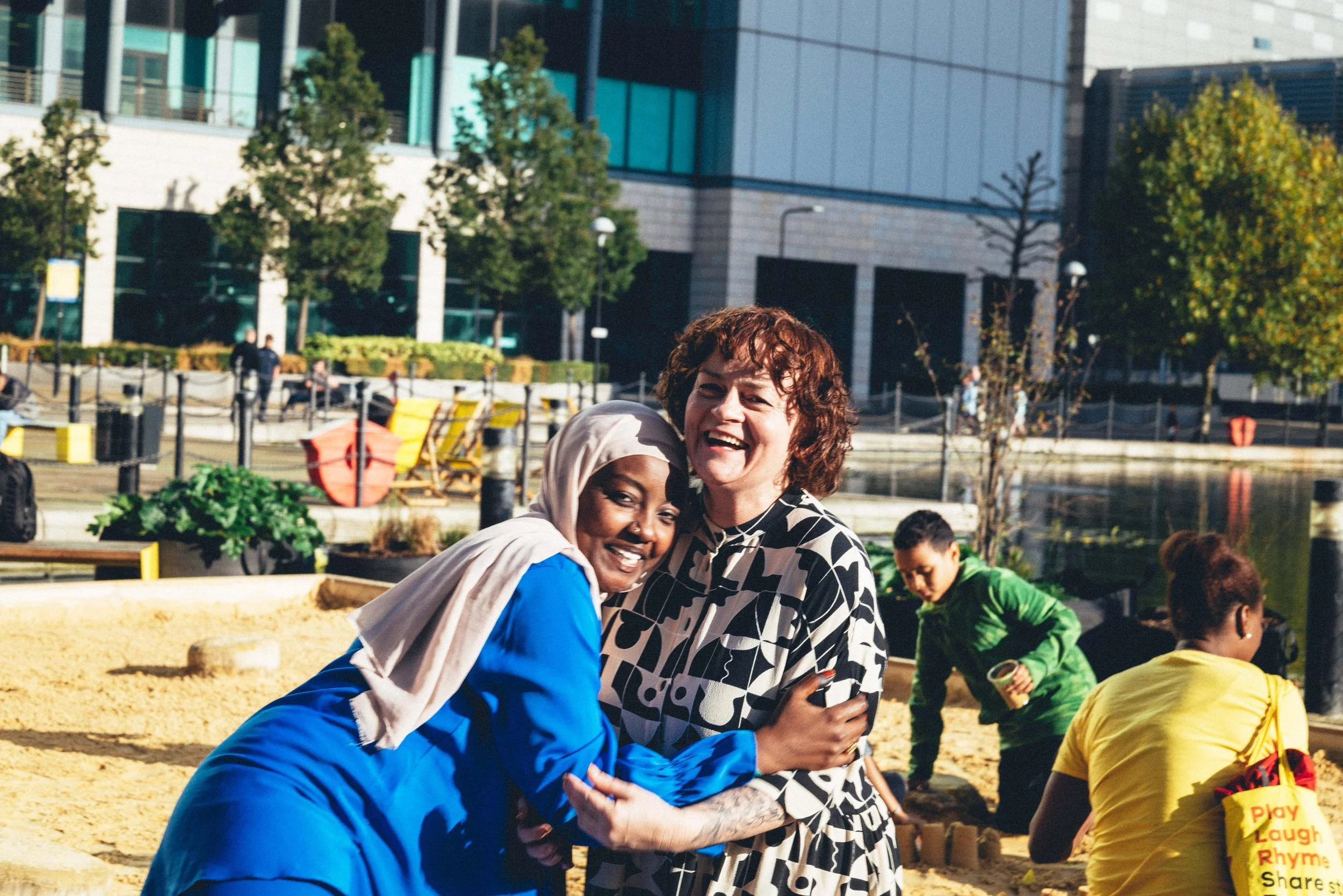 Two women smiling and hugging in a park near a body of water, with trees and modern buildings in the background, and other people working on a project or playing.