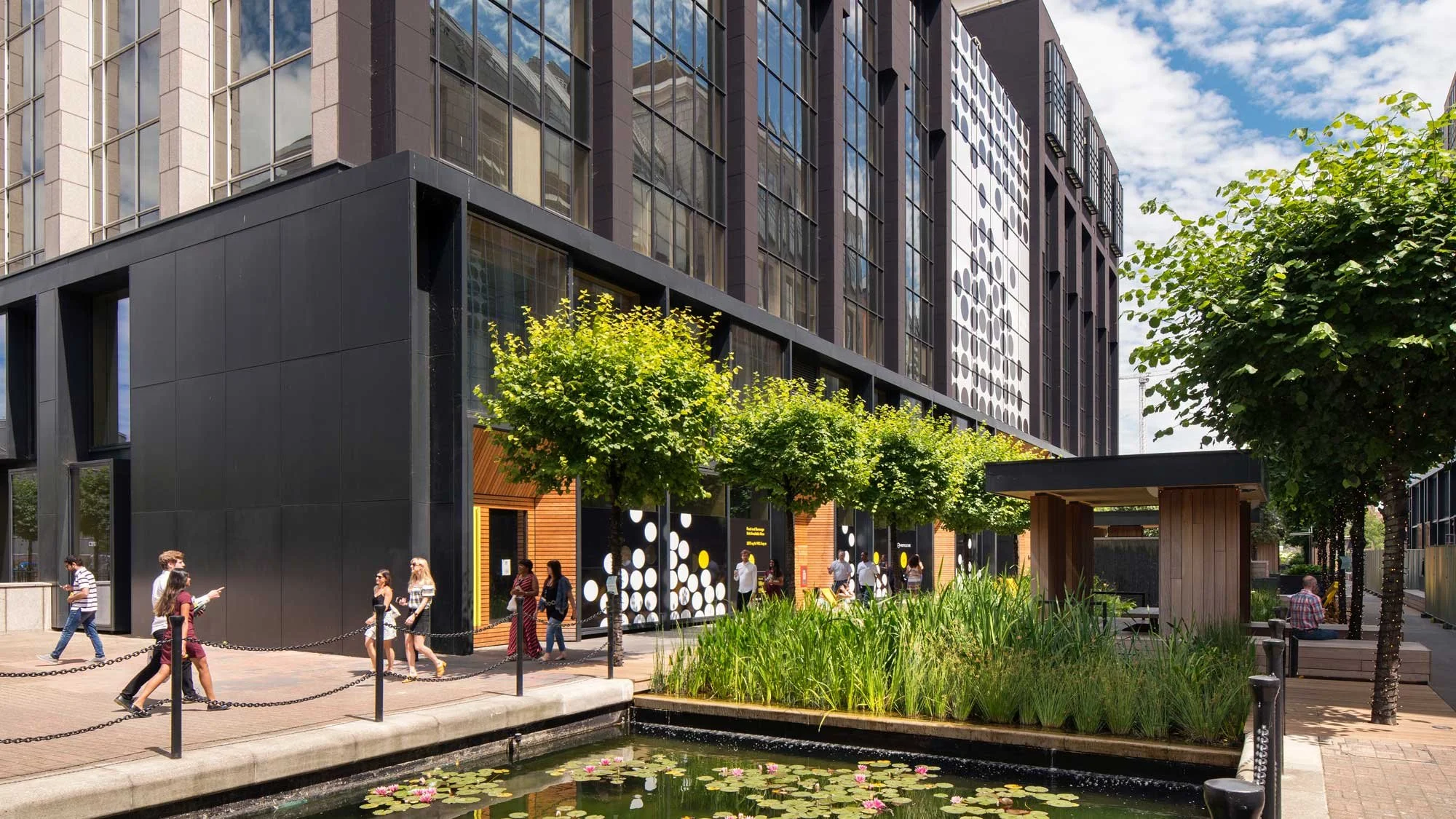 View of a modern building with glass windows and black exterior, with people walking outside near a pond with lily pads, trees, and a small wooden shelter under a partly cloudy sky.