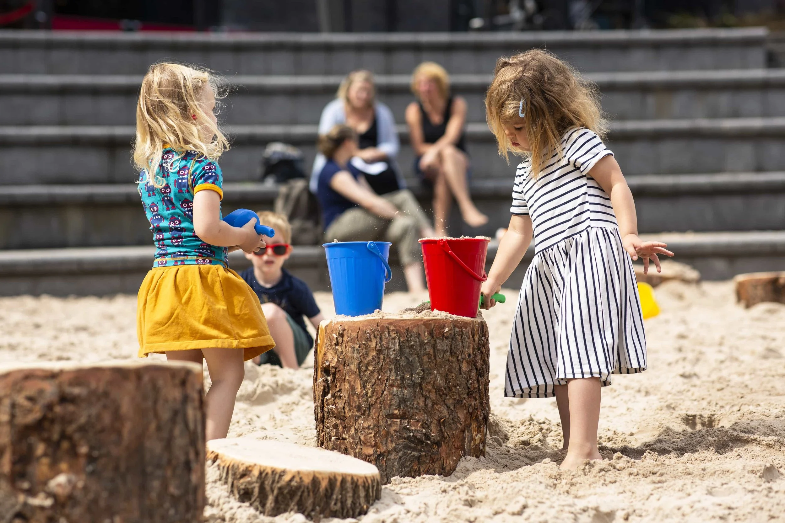 Two young girls playing with toy shovels and buckets on a sandy surface, with three women seated on steps in the background.