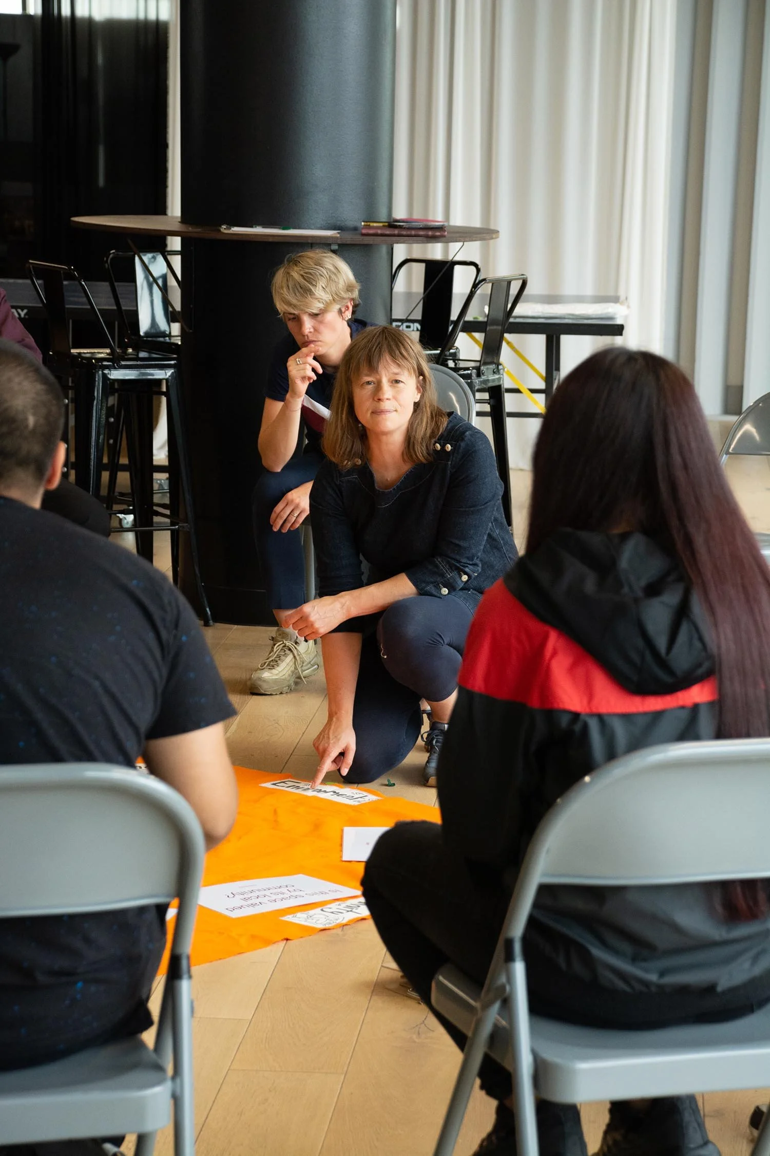 A group of people participating in a discussion or activity, sitting on chairs around a large orange poster on the floor in a room with large windows and curtains.