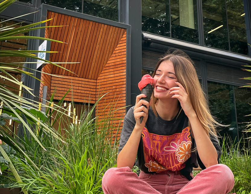 A young woman with long hair, wearing a black t-shirt with colorful graphics and pink pants, sitting outdoors on grass, eating ice cream and smiling.
