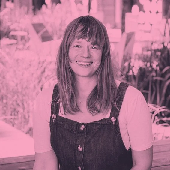 A woman with shoulder-length hair and bangs smiling, wearing a white shirt and a dark overall dress, sitting in an outdoor setting with plants and flowers in the background.