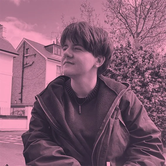 A young person with short dark hair sitting outdoors near buildings and trees, with a pink filter applied to the photo.