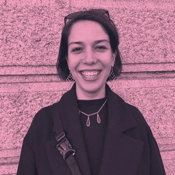 A woman with short dark hair, wearing earrings, a necklace, and a black jacket, smiling in front of a textured wall with a pink filter.