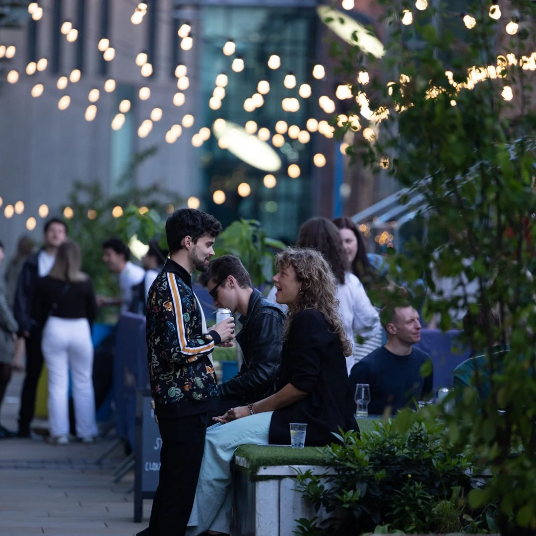 People socializing outdoors at an evening gathering with string lights overhead, standing and sitting near greenery.